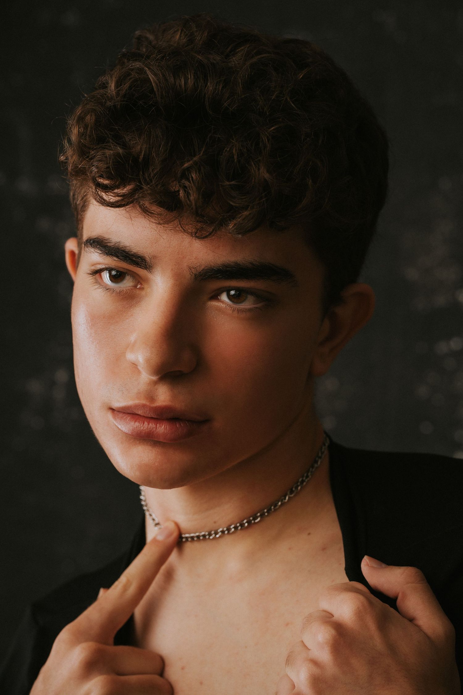 Young male model with curly hair wearing silver chain necklace against dark background.