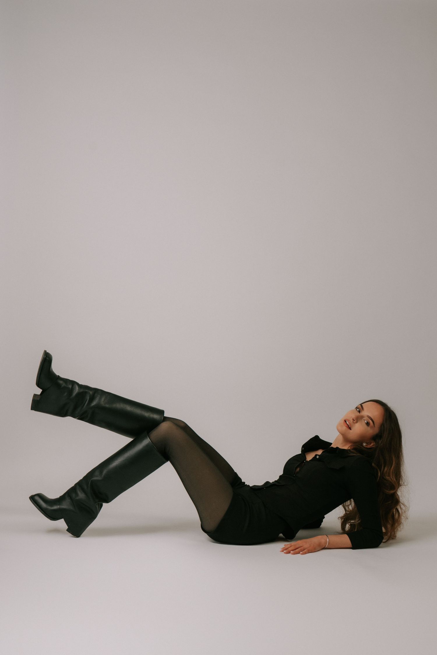 Brunette woman in black outfit and tall leather boots lying on studio floor looking up.