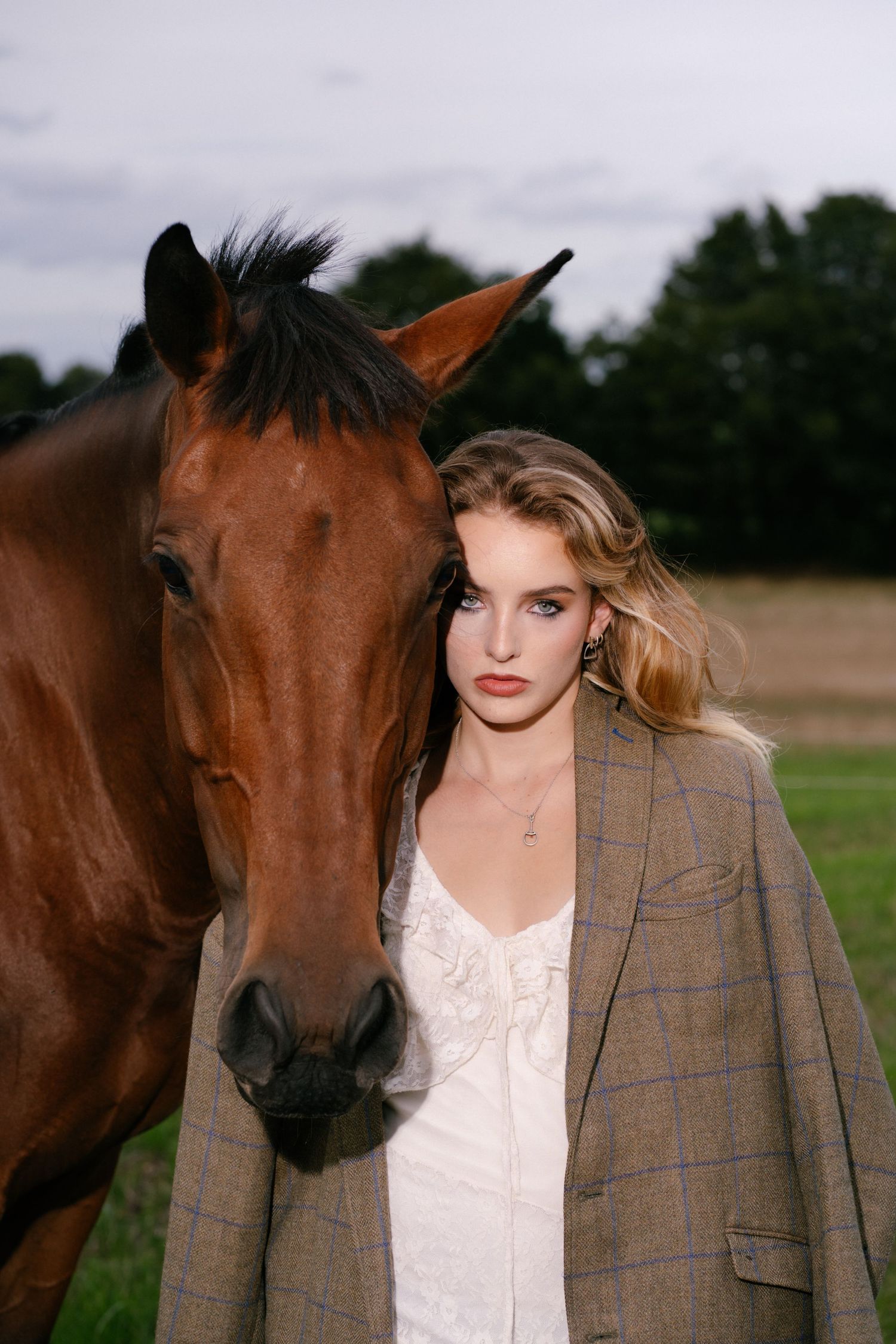 Young blonde woman standing beside a bay horse wearing a white dress and oversized plaid blazer.