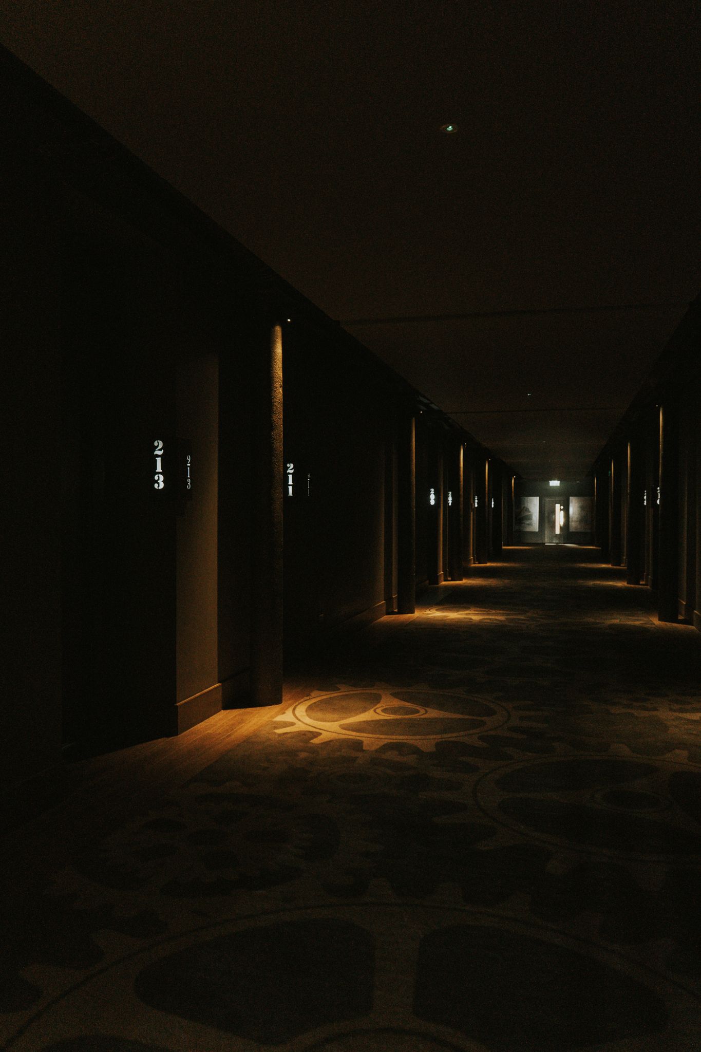 Dark moody hotel corridor with patterned carpet and subtle warm lighting along room doors.