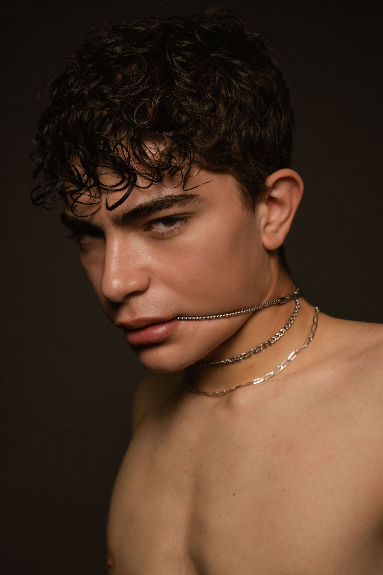 Close-up portrait of male model with curly hair wearing layered silver chains on dark background.