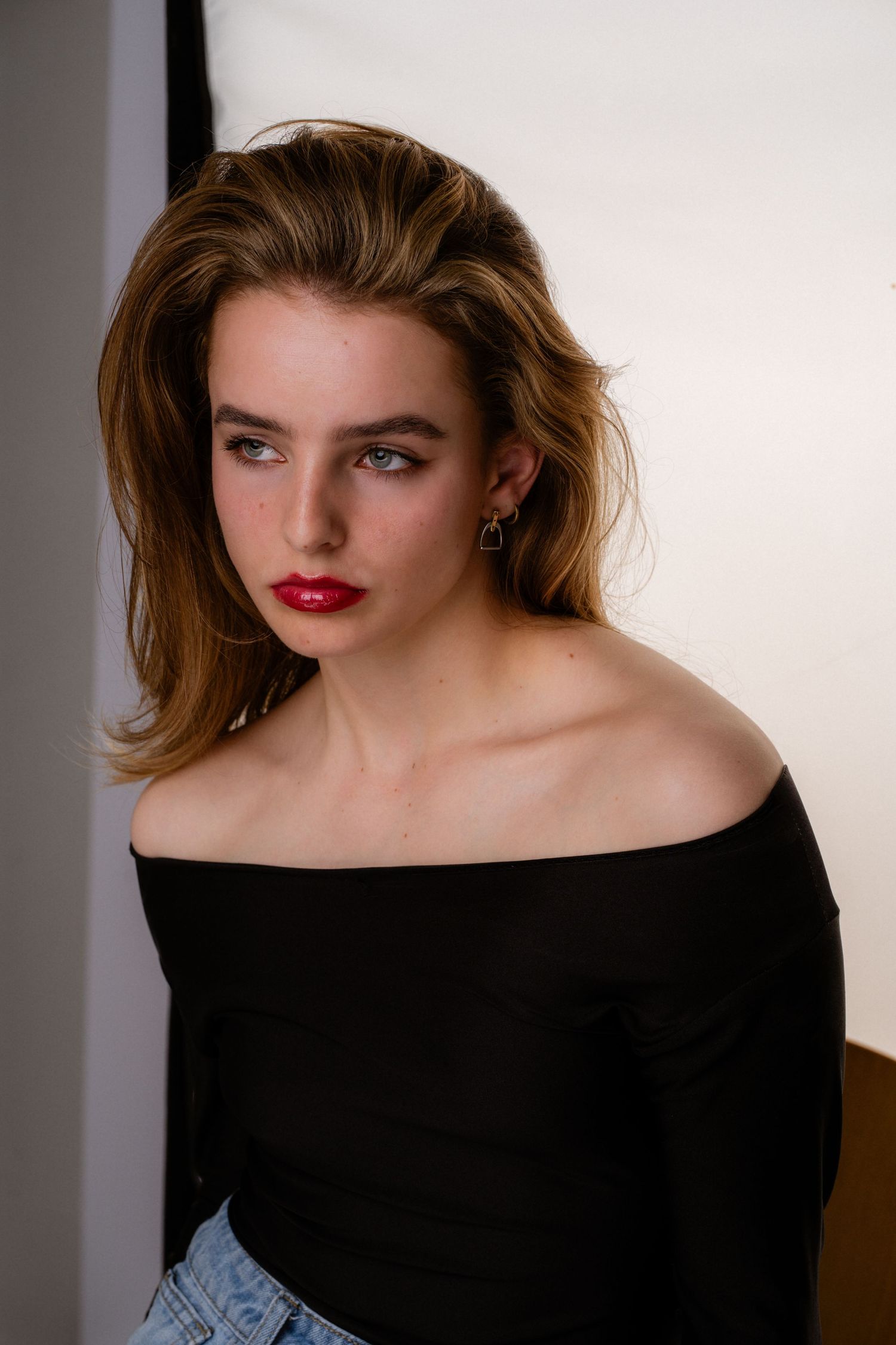 Young woman in off-shoulder black top with red lips and earring poses in a studio setting.