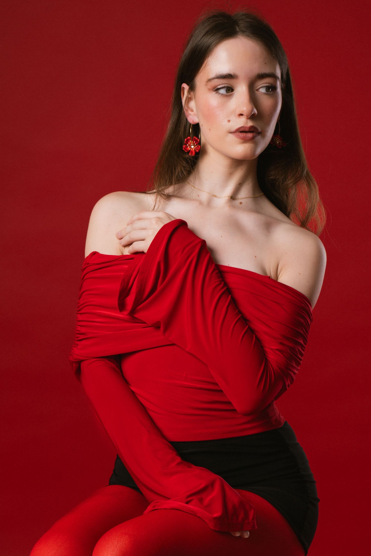 Elegant woman in red off-shoulder draped top with matching earrings poses on red background.