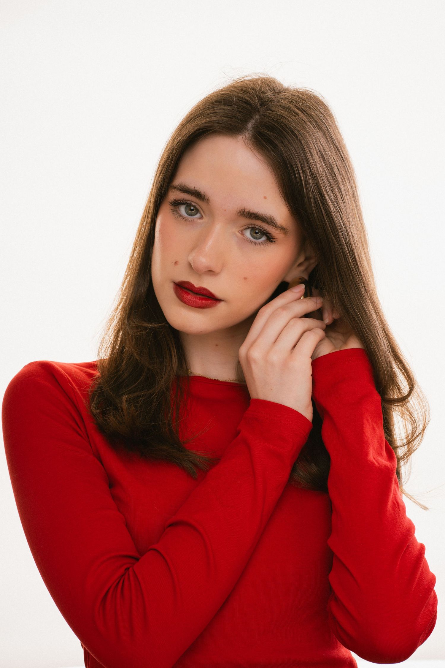 Young brunette woman in red long-sleeve top with red lips poses against a white background.