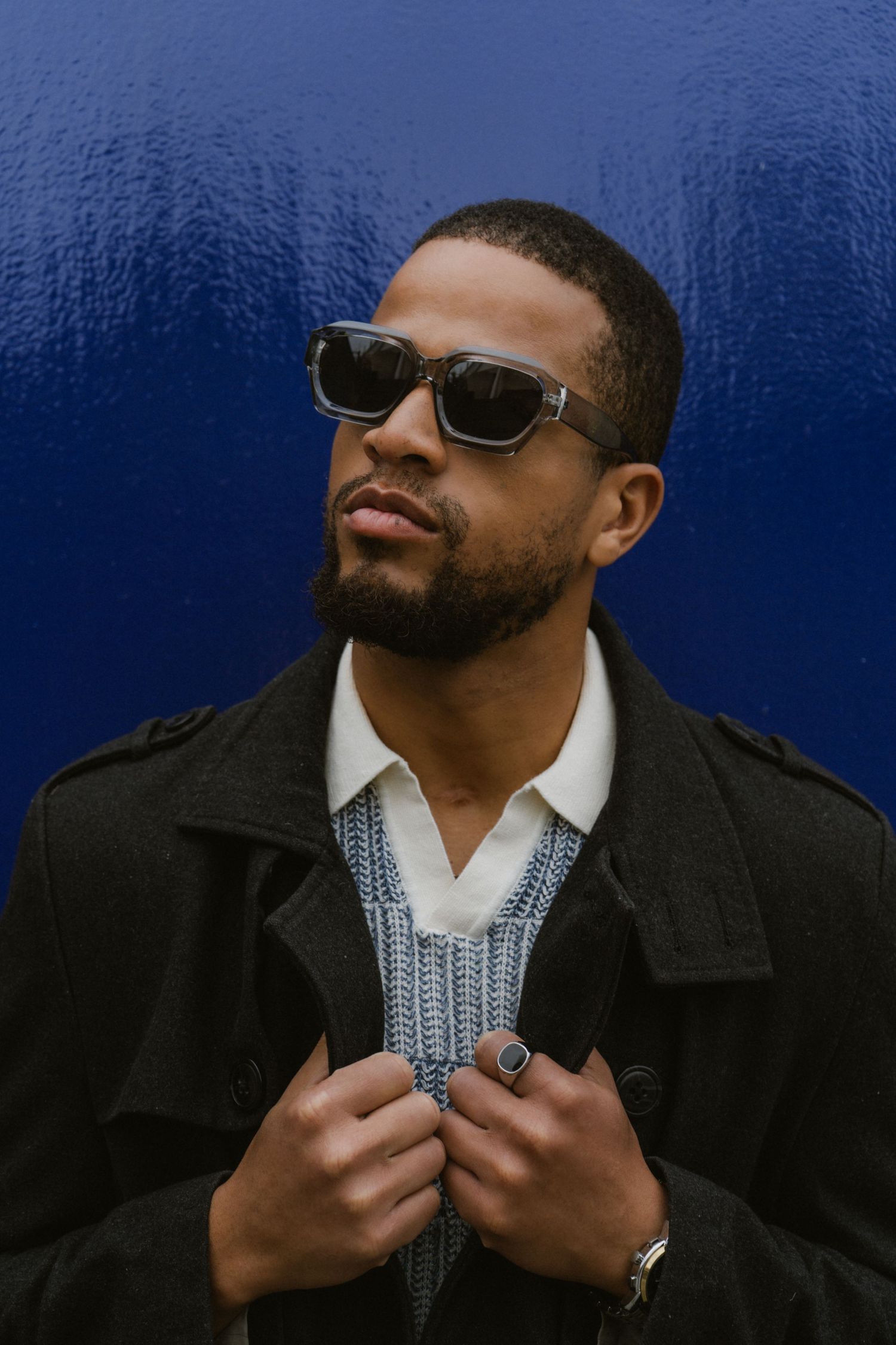 Stylish man in sunglasses and black trench coat posing against a vibrant blue wall.