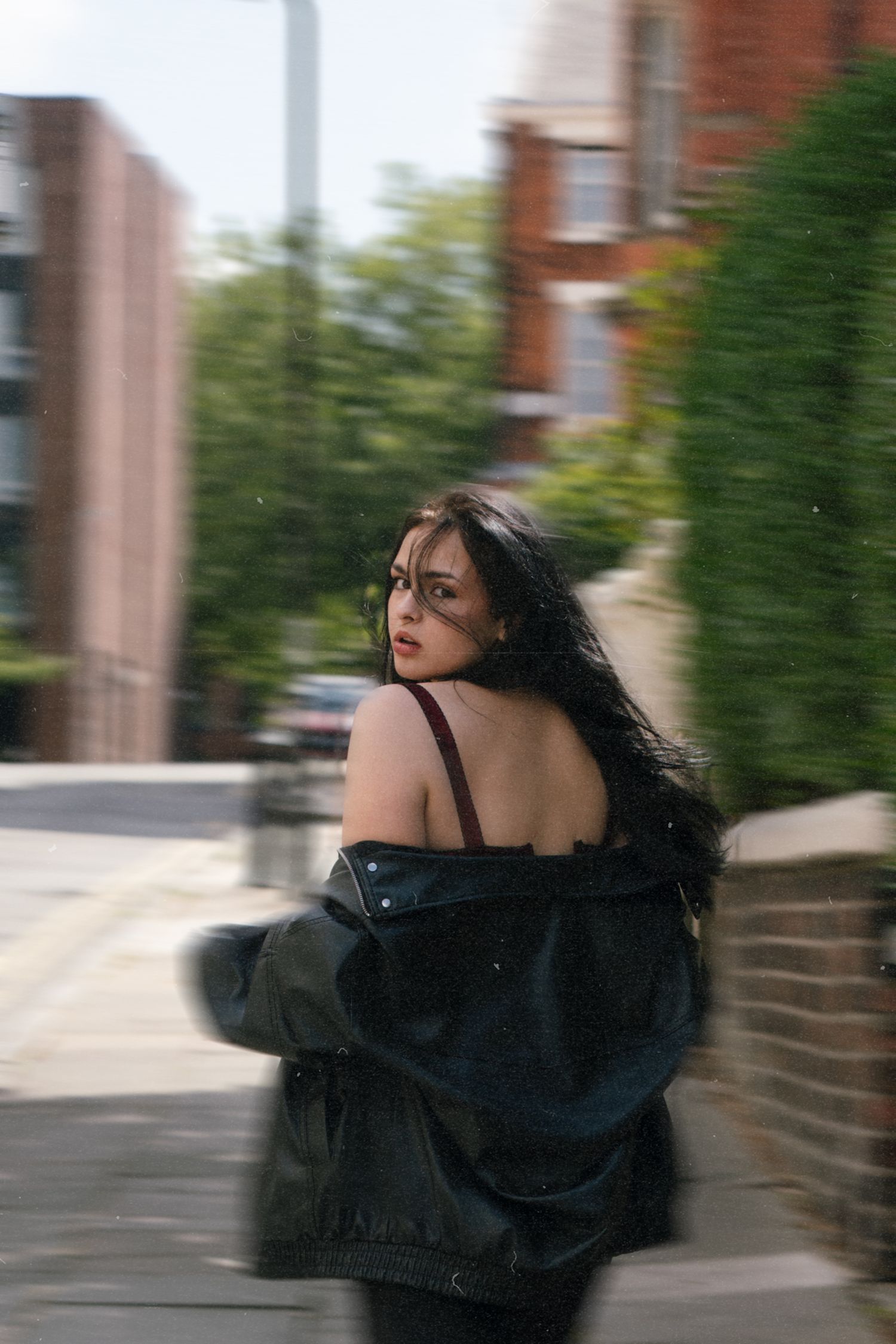 Young woman with long black hair in leather jacket glances back on urban street sidewalk.