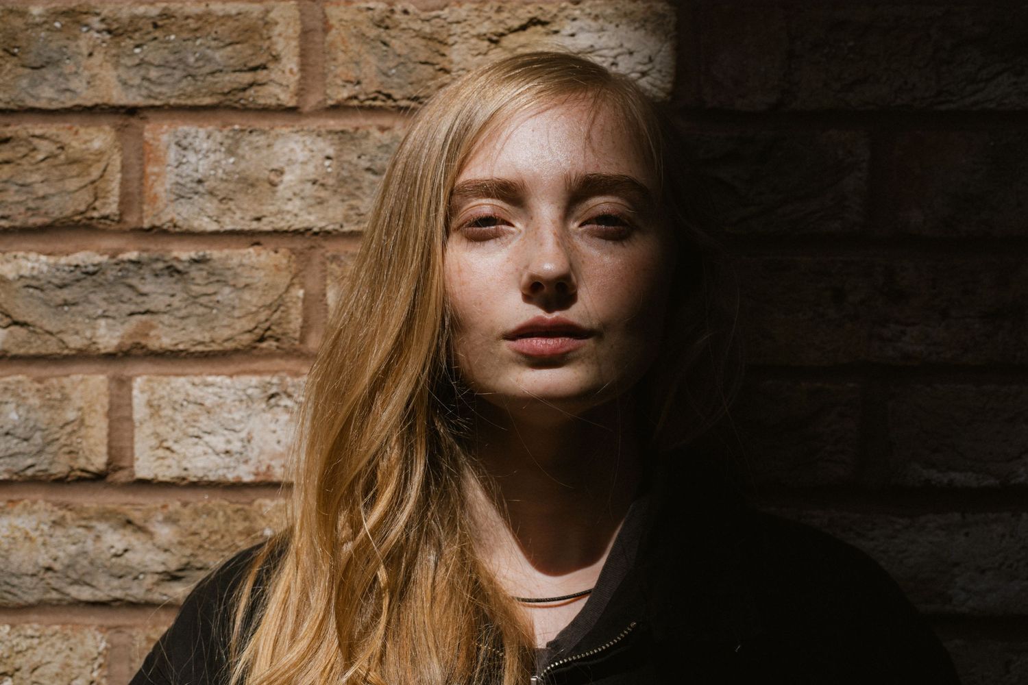 Young woman with long blonde hair leaning against a brick wall in dramatic natural lighting.
