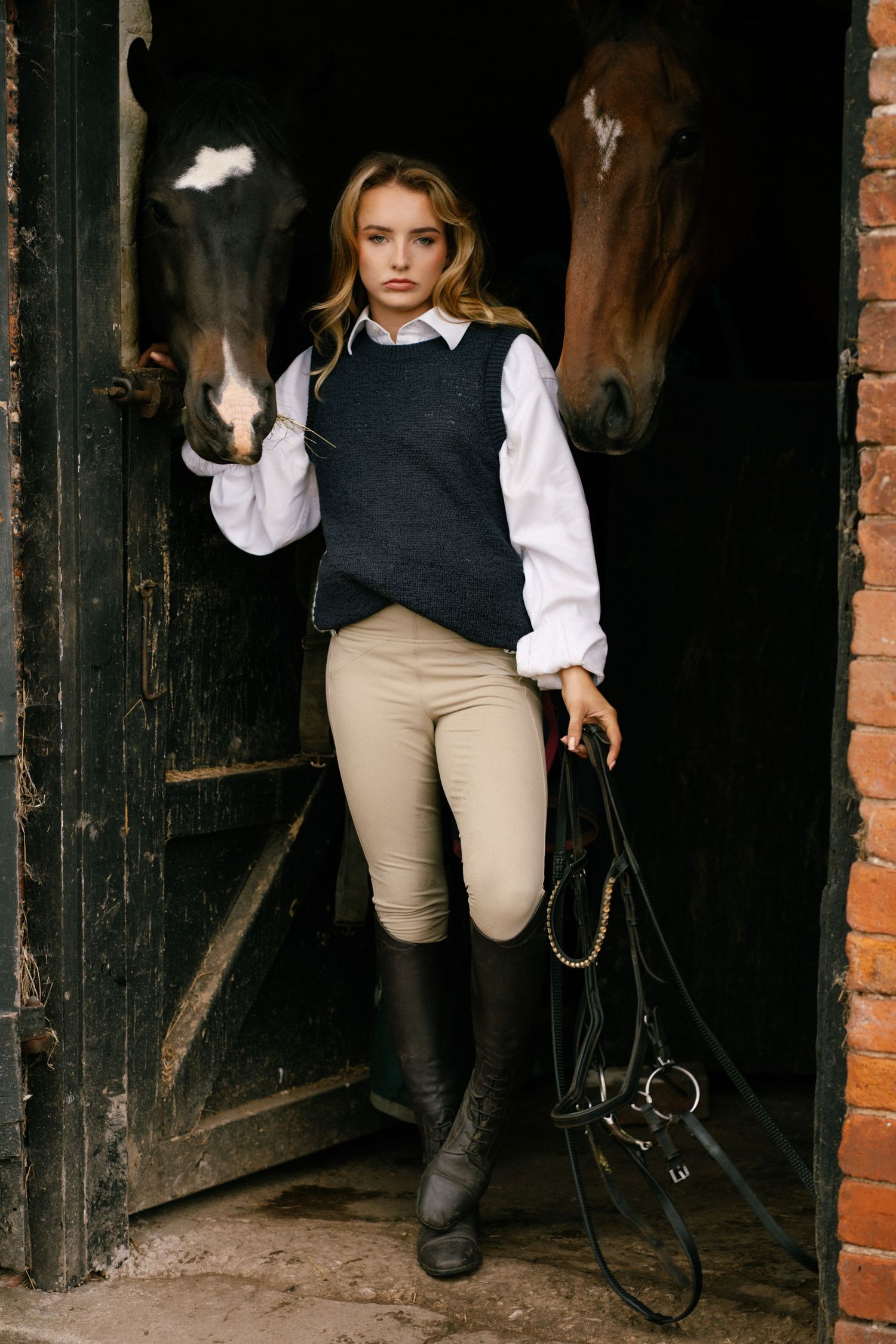 Young woman in equestrian attire holding reins beside two horses at stable doorway.