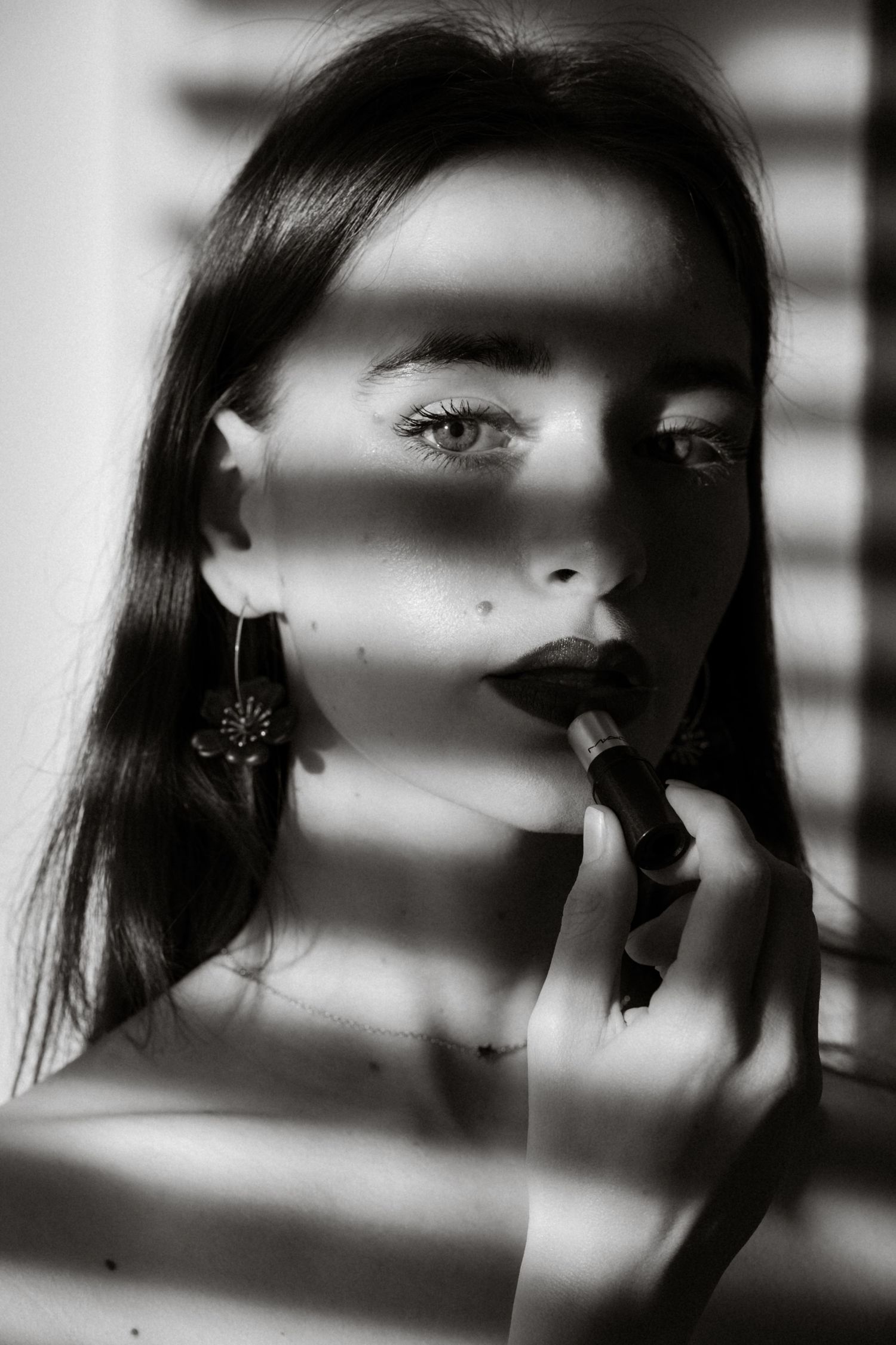 Black and white close-up portrait of a woman applying lipstick with shadow stripes across her face.