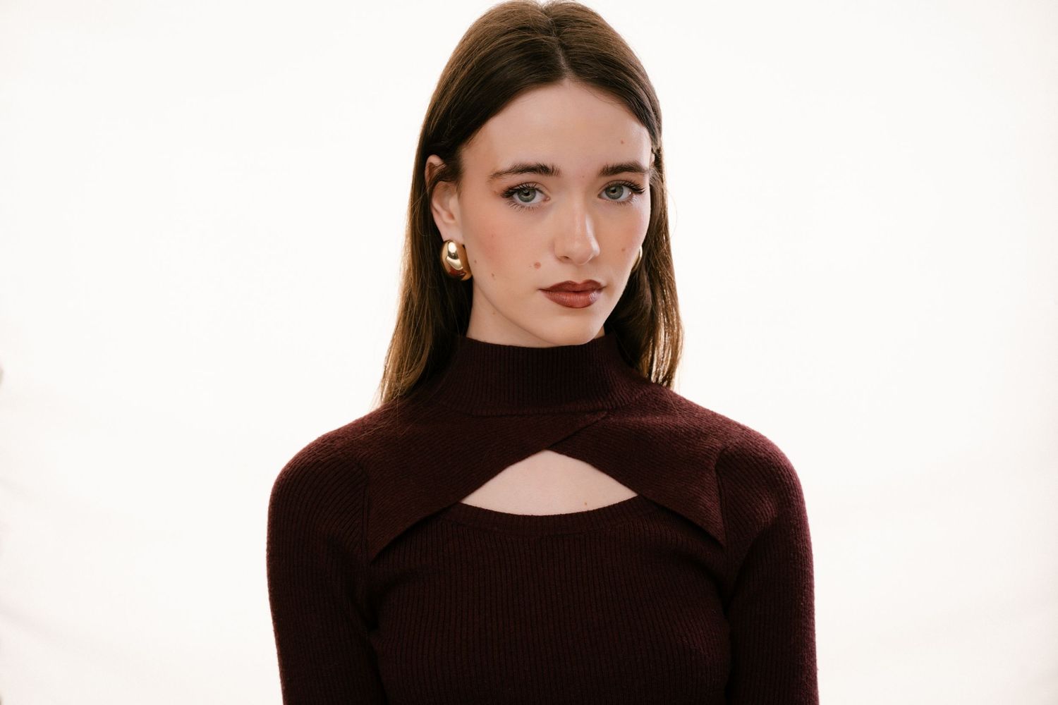 Young woman in burgundy cutout turtleneck top with gold earrings poses on white background.