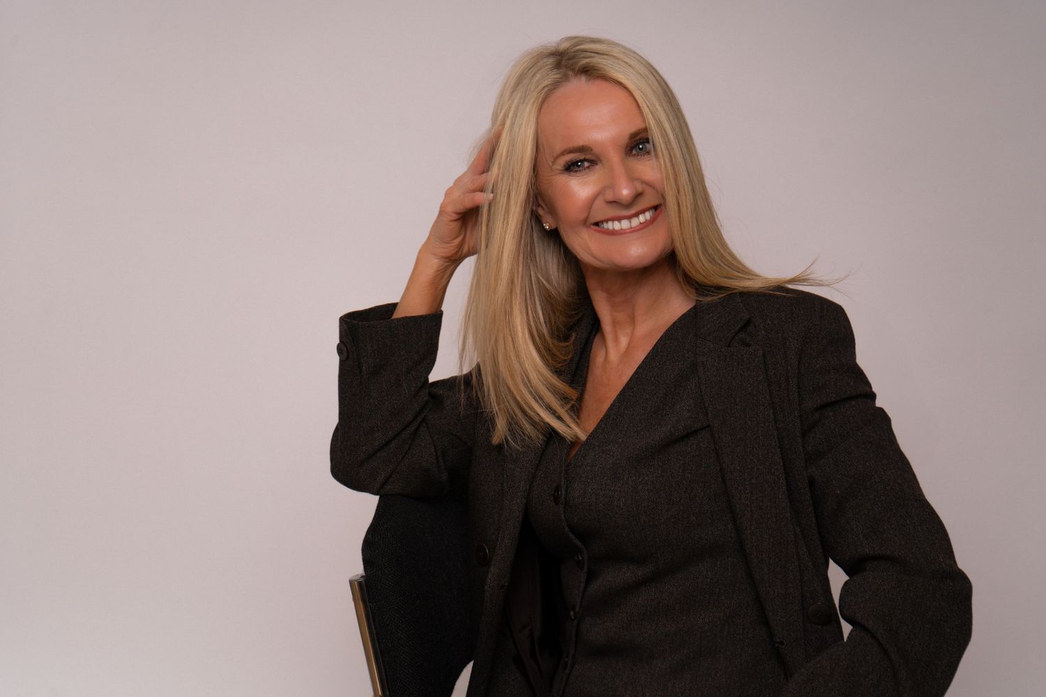 Smiling blonde woman in dark blazer sitting casually with hand in hair in bright studio.
