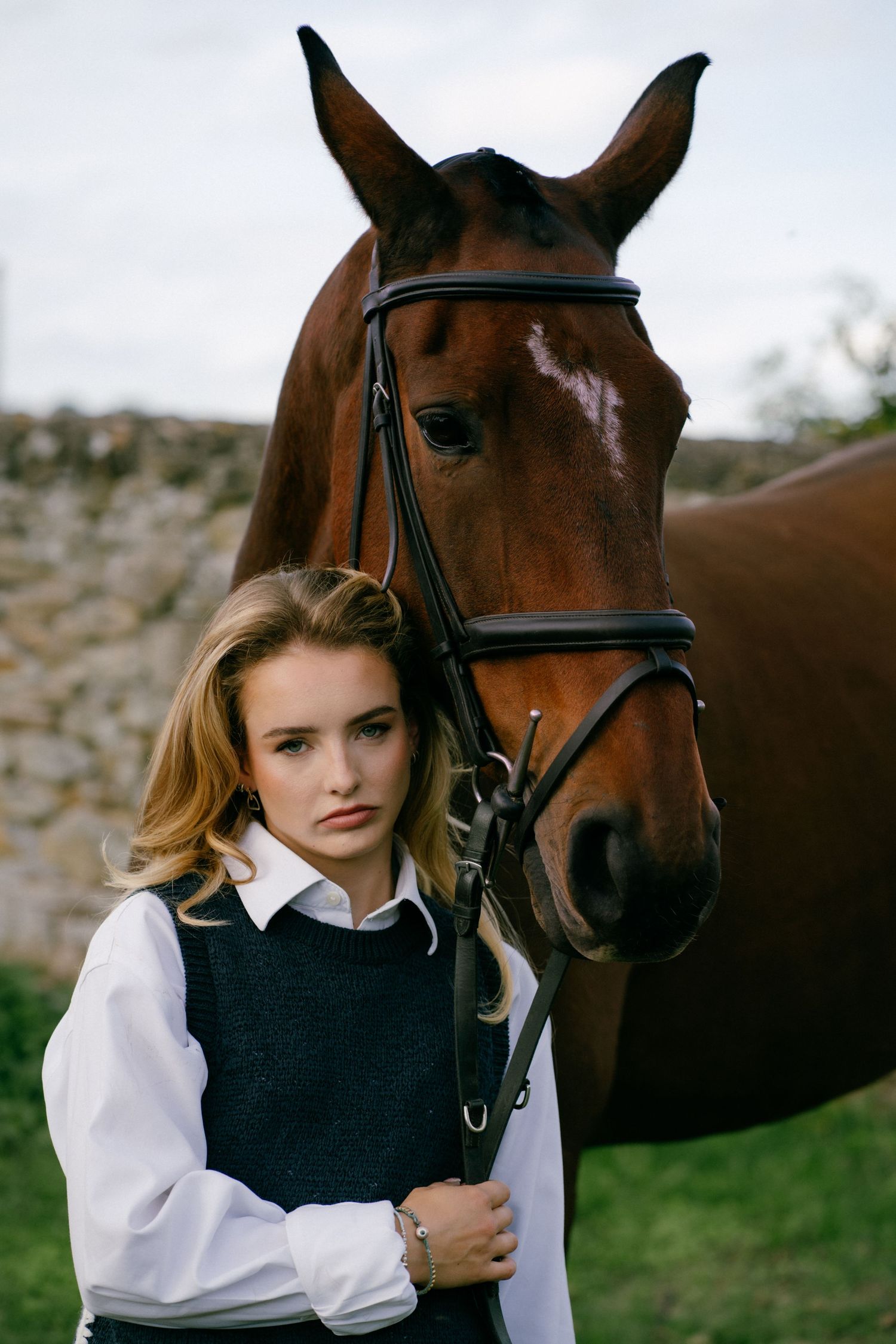 Young blonde woman in white shirt and black vest standing beside a bay horse wearing a bridle outdoors.