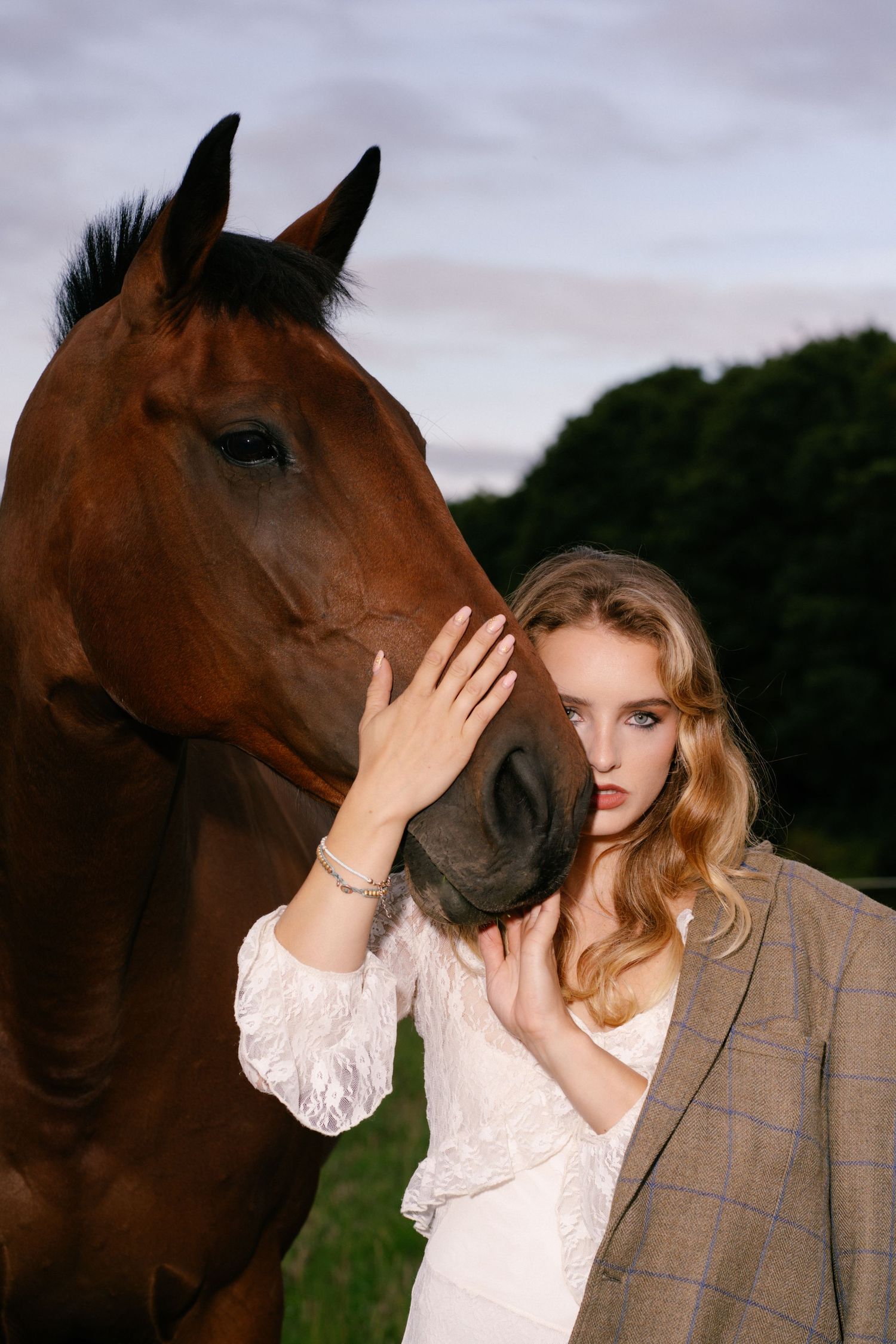 Young blonde woman in white dress and blazer gently touching a brown horse's face outdoors at dusk.