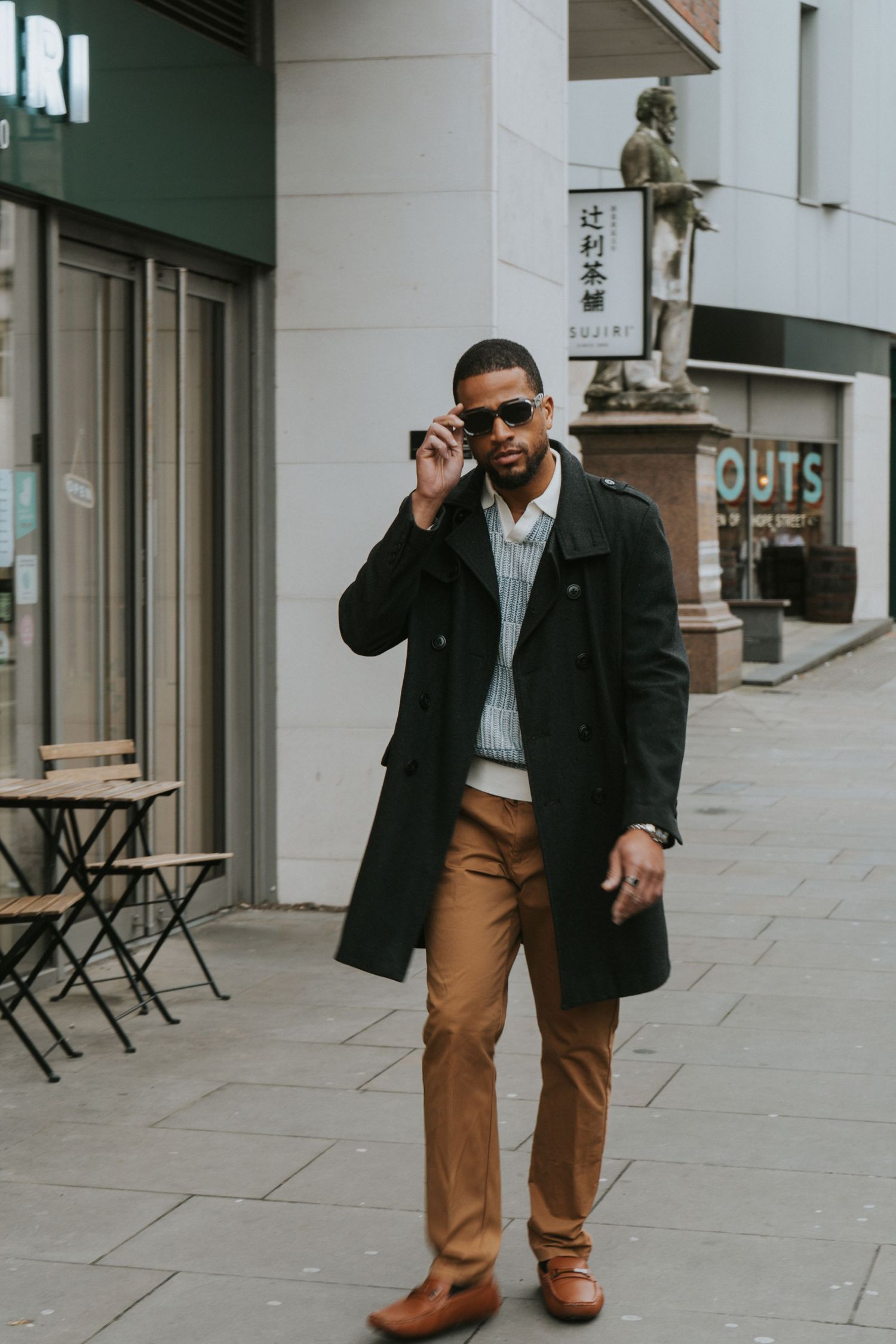 Stylish man in black overcoat and tan trousers talking on phone on a city street.