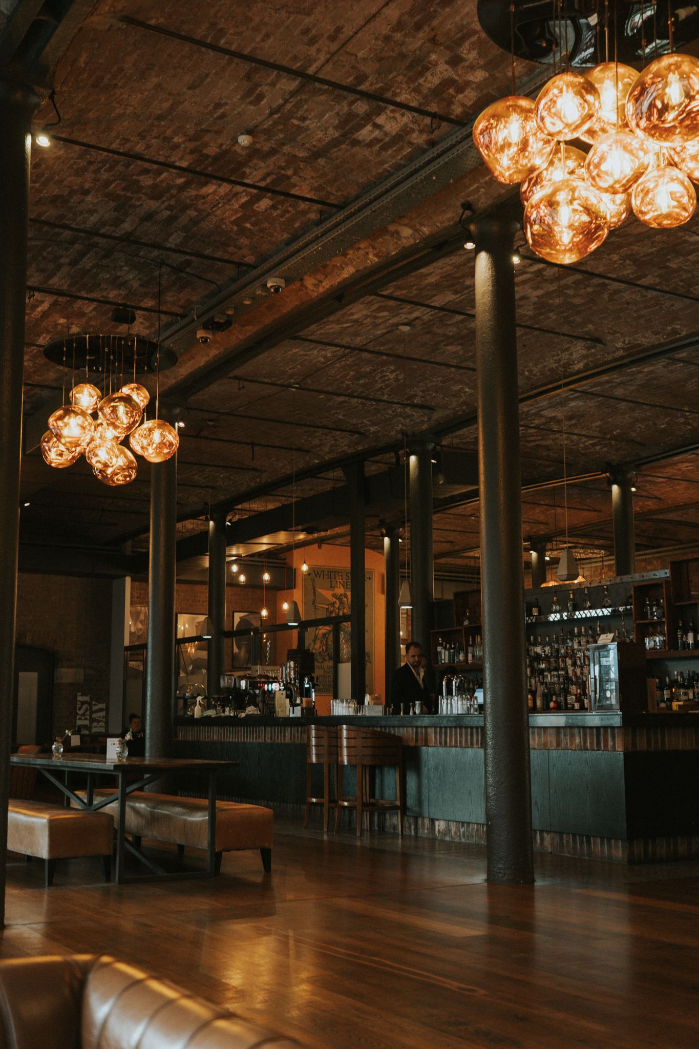 Dimly lit hotel bar with exposed brick ceiling, amber globe chandeliers, and wood floors.