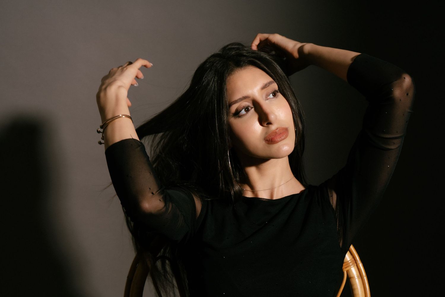 Brunette woman in black outfit with hands in hair, making pouty expression in dark studio.