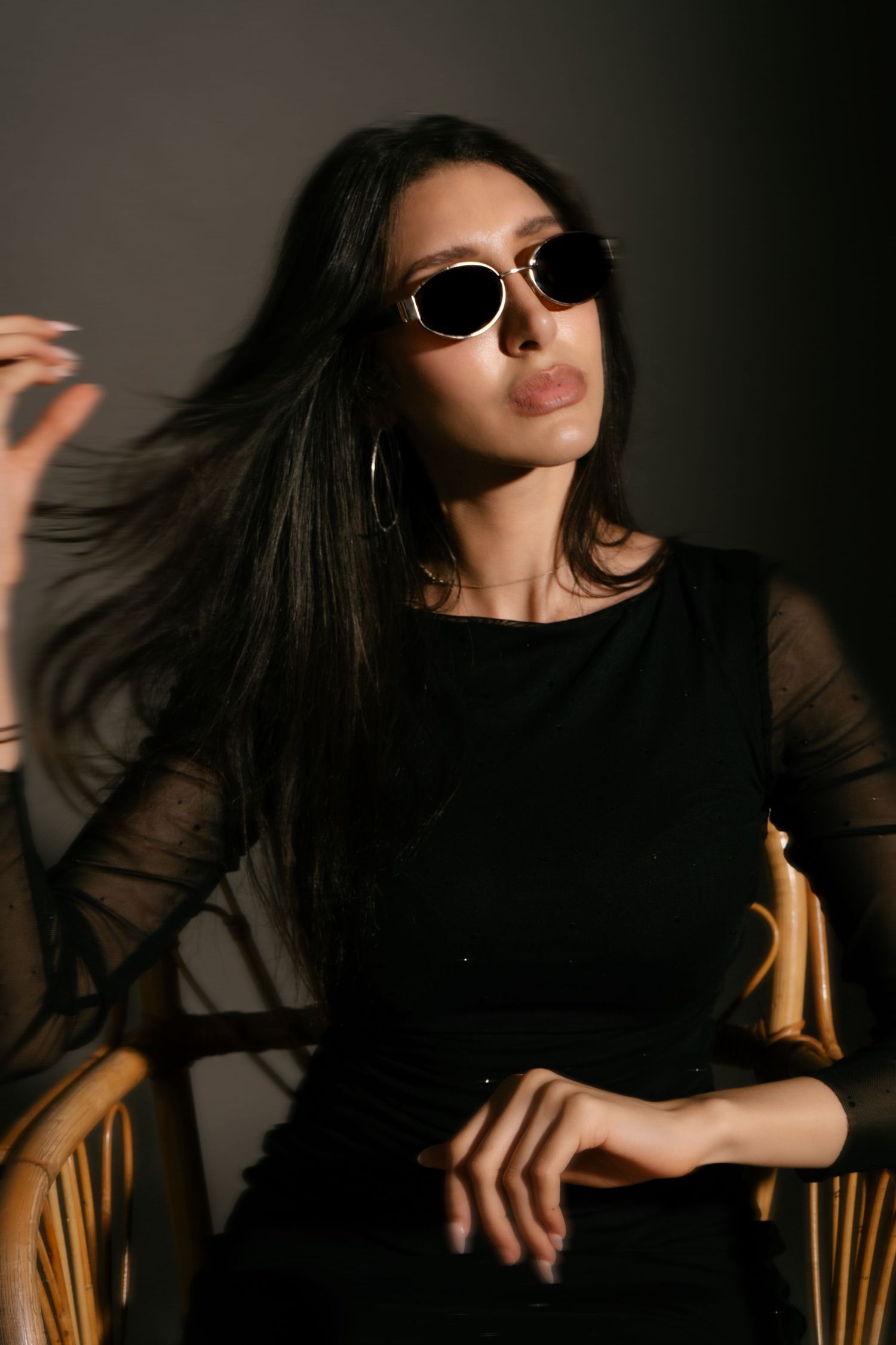 Stylish young woman in black outfit and round sunglasses sitting in a wicker chair on dark background.