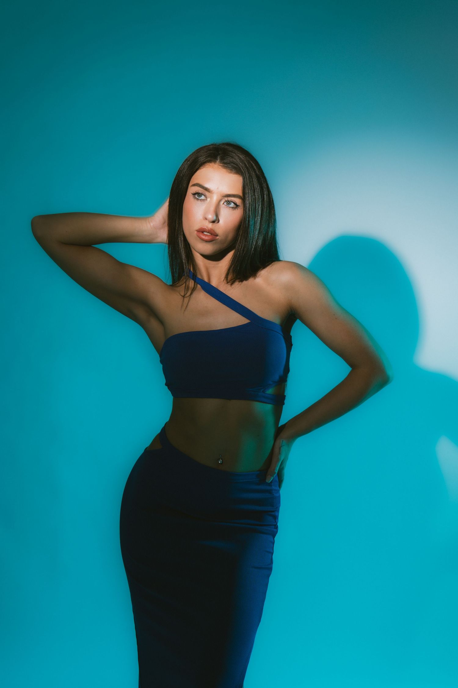 Woman in blue one-shoulder crop top and skirt posing against bright turquoise background in studio.