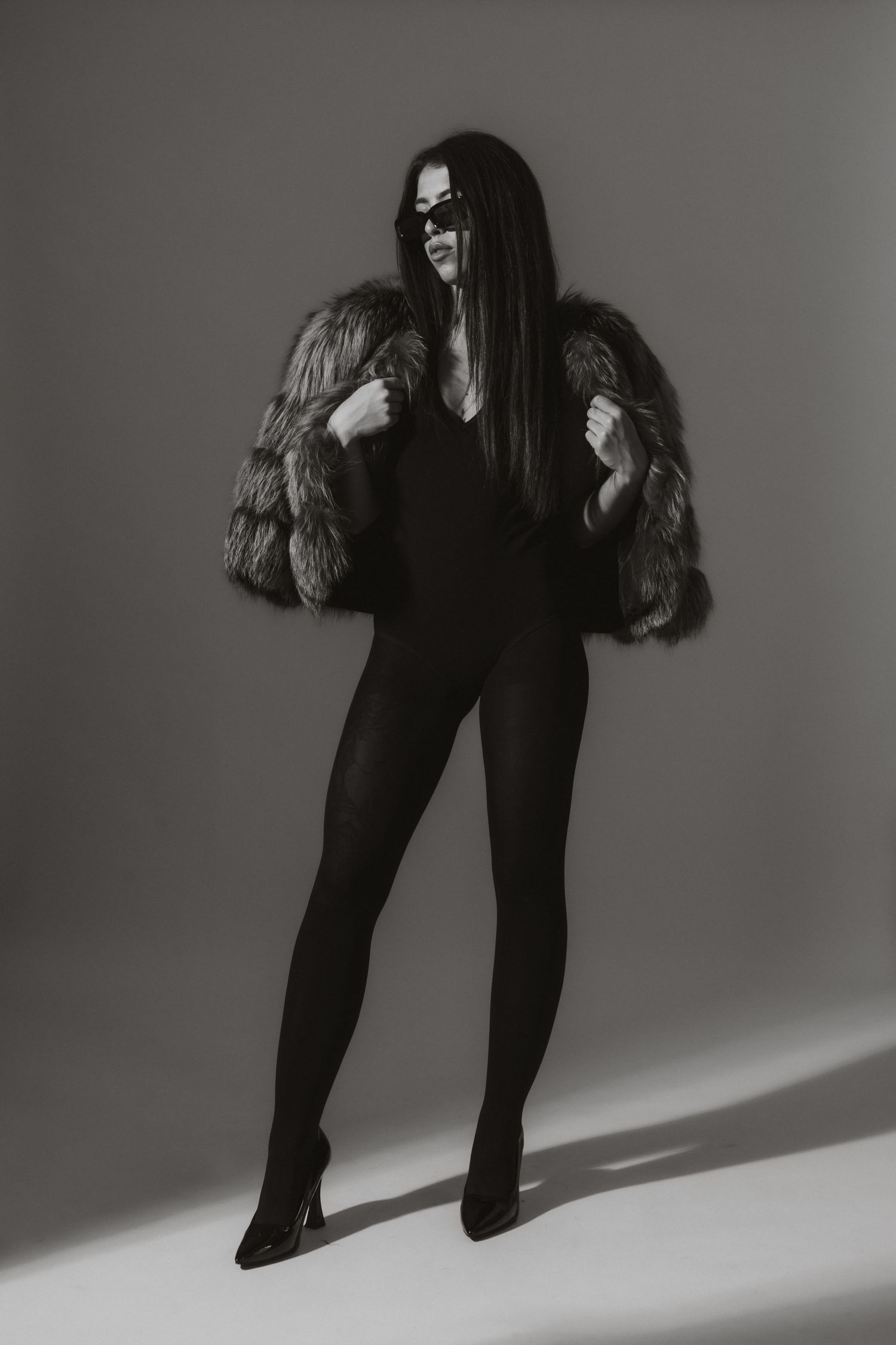 Full-length black and white portrait of woman in fur coat and black bodysuit with sunglasses.