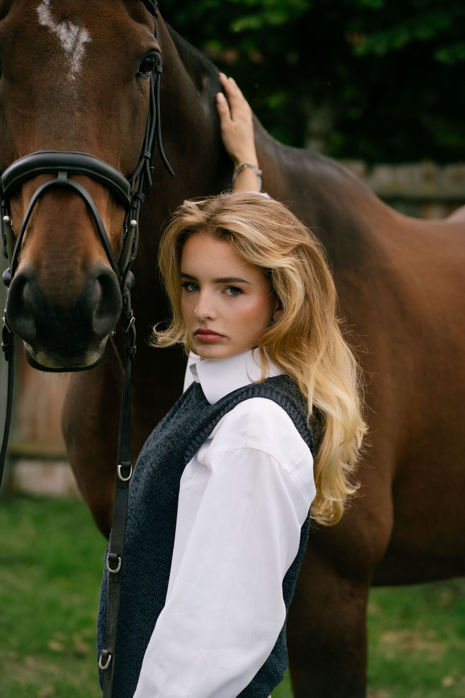 Young blonde woman in white blouse and gray vest poses beside a brown horse with bridle outdoors.