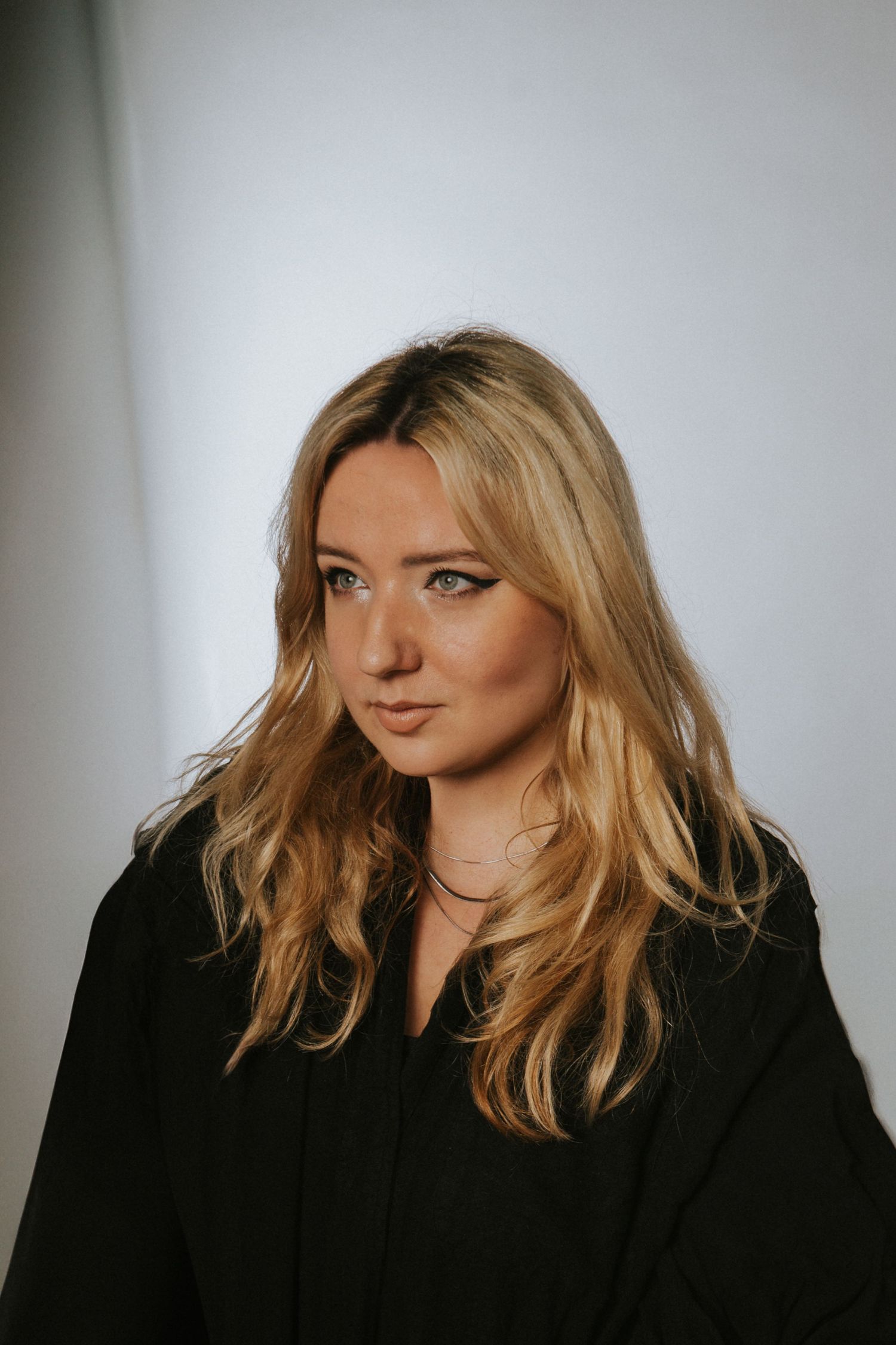 Blonde woman in black top with soft waves posing against light gray background.