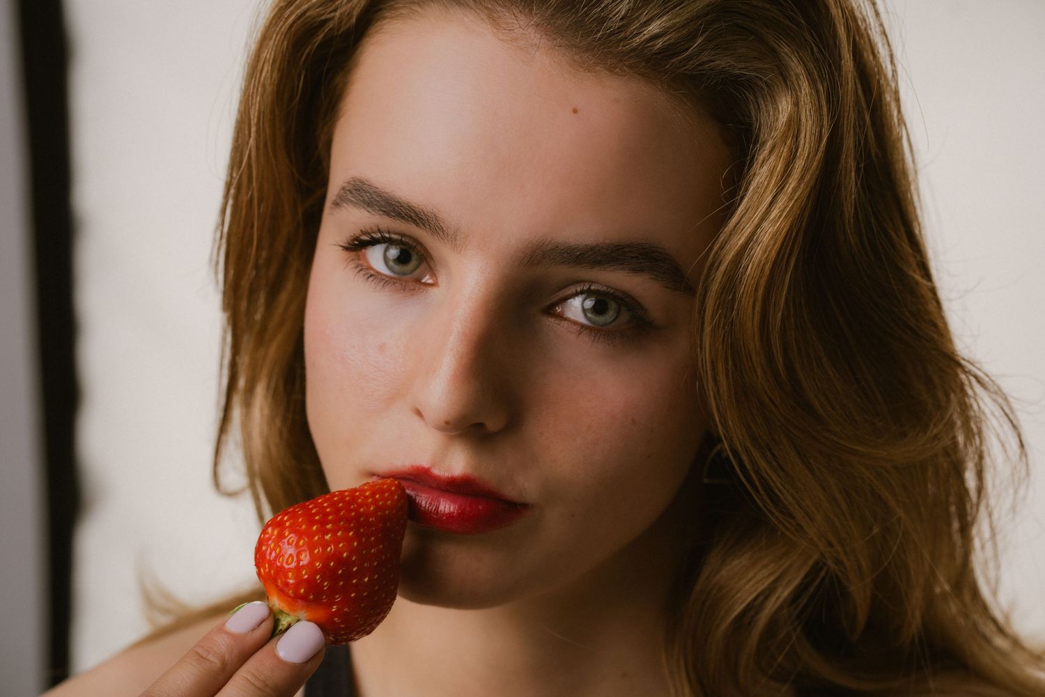 Young woman with auburn hair and red lips holding a fresh strawberry near her mouth.