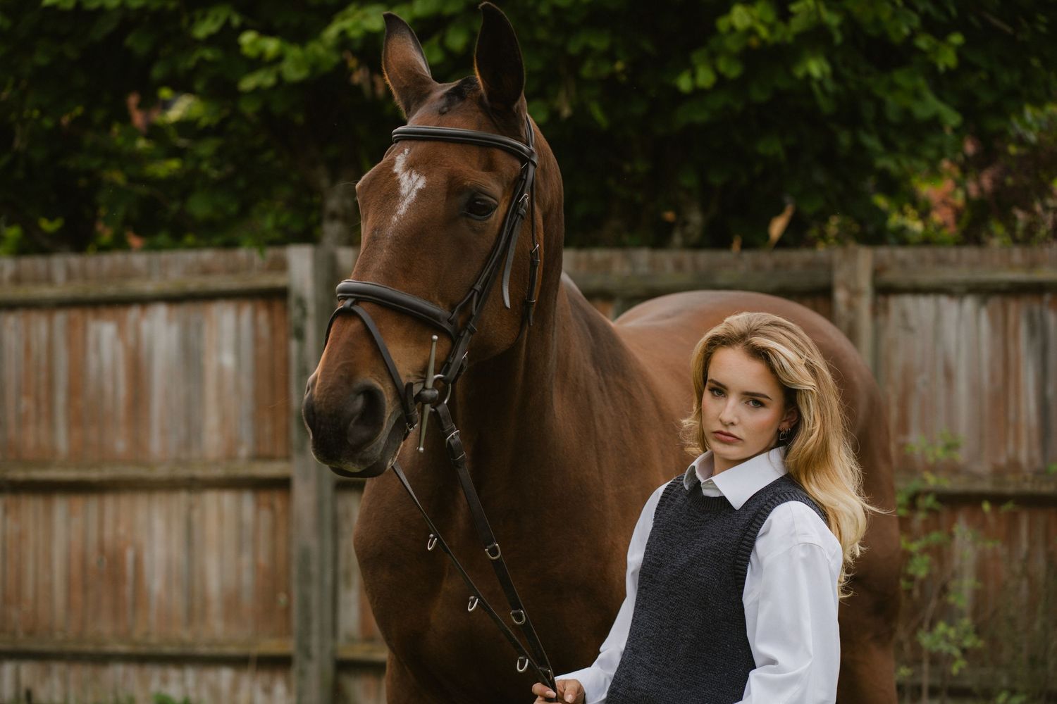 Young blonde woman in equestrian attire standing beside a chestnut horse near a wooden fence.