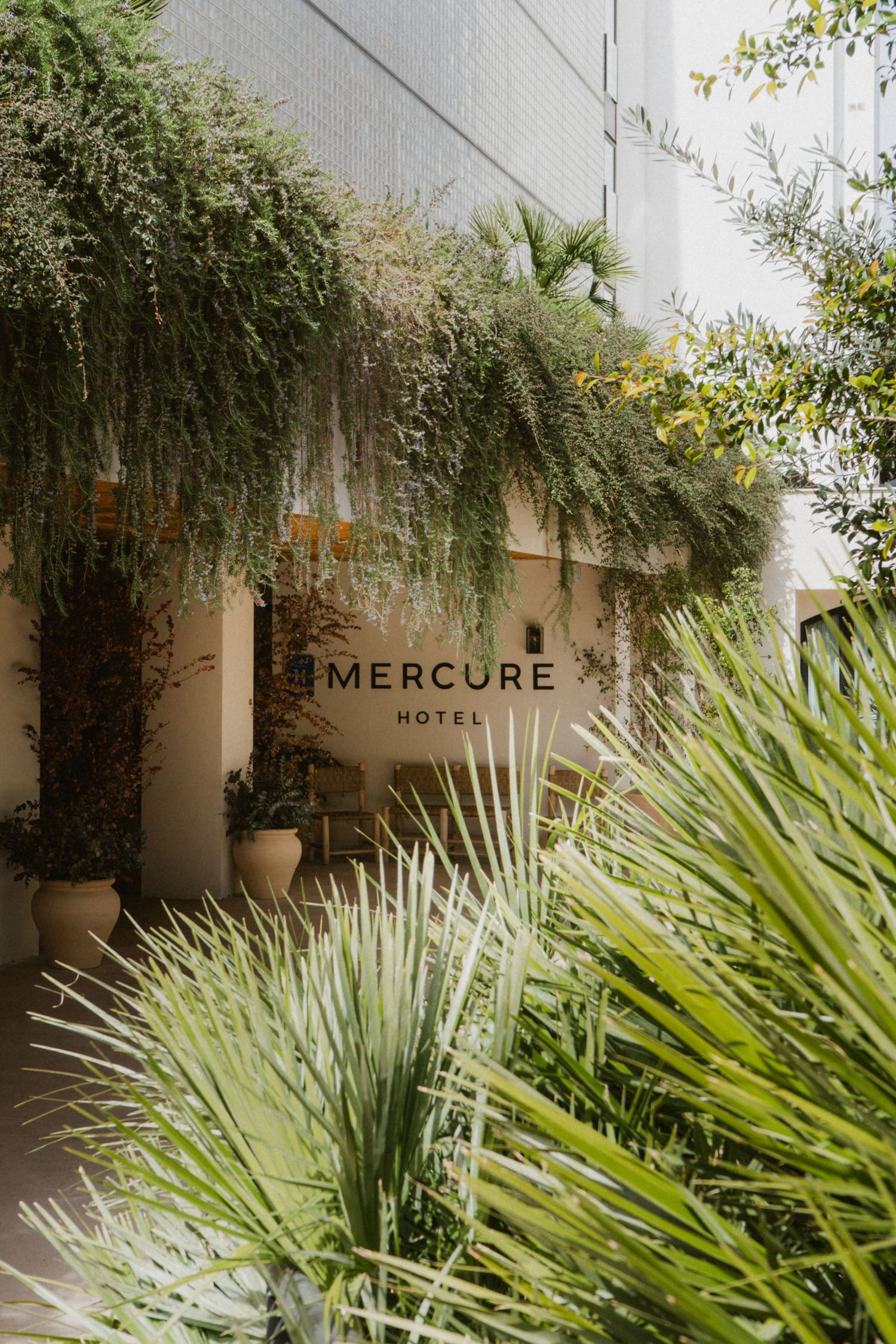 Lush green entrance of Mercure Hotel surrounded by tropical plants, palm fronds, and hanging foliage on a bright day.