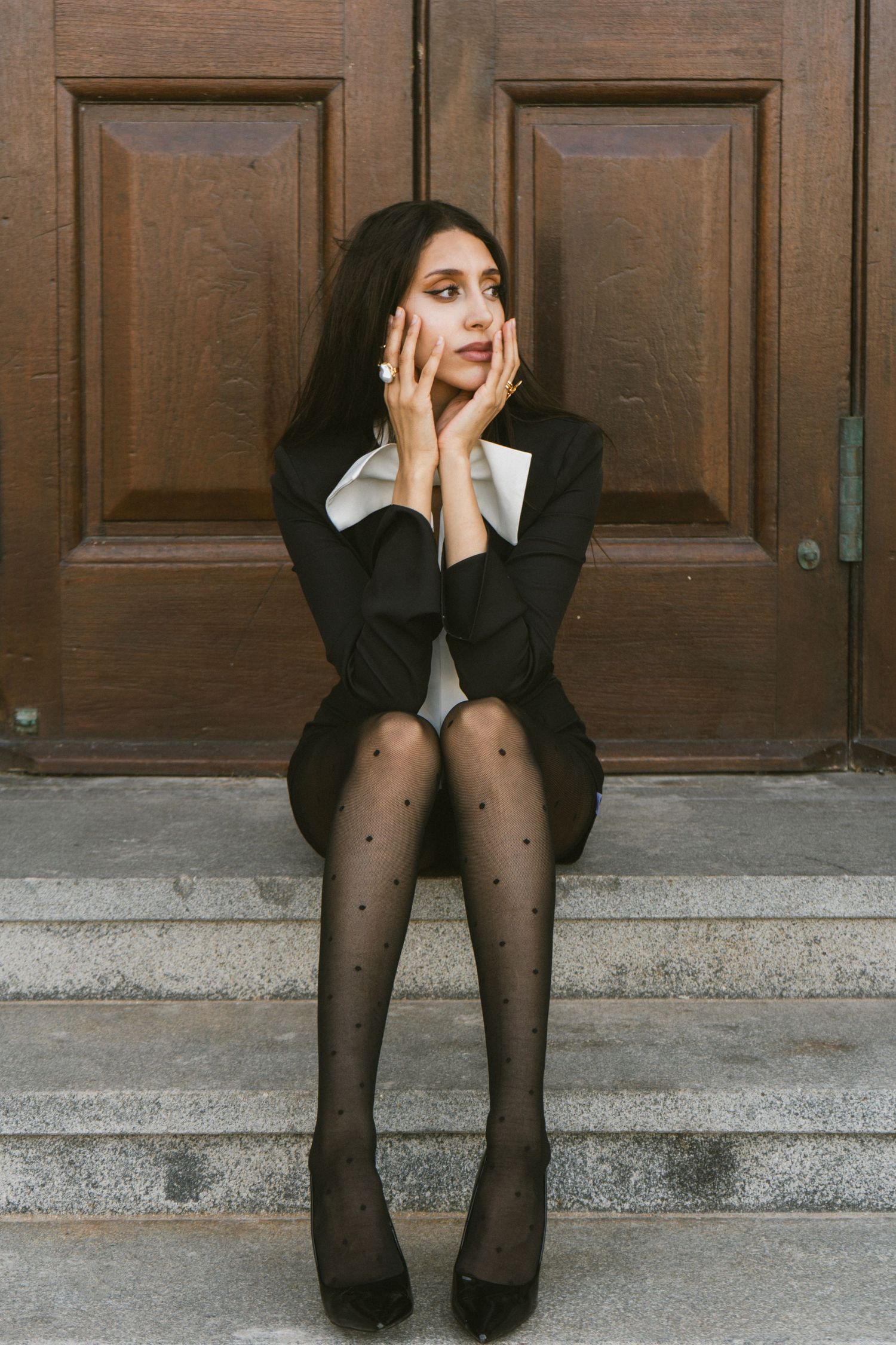 Young woman in black blazer and polka dot tights sits on stone steps before a wooden door, hands framing her face.