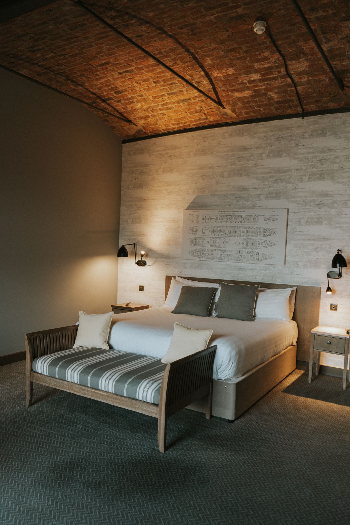 Elegant hotel bedroom with exposed brick ceiling, large bed, striped bench, and warm lighting.