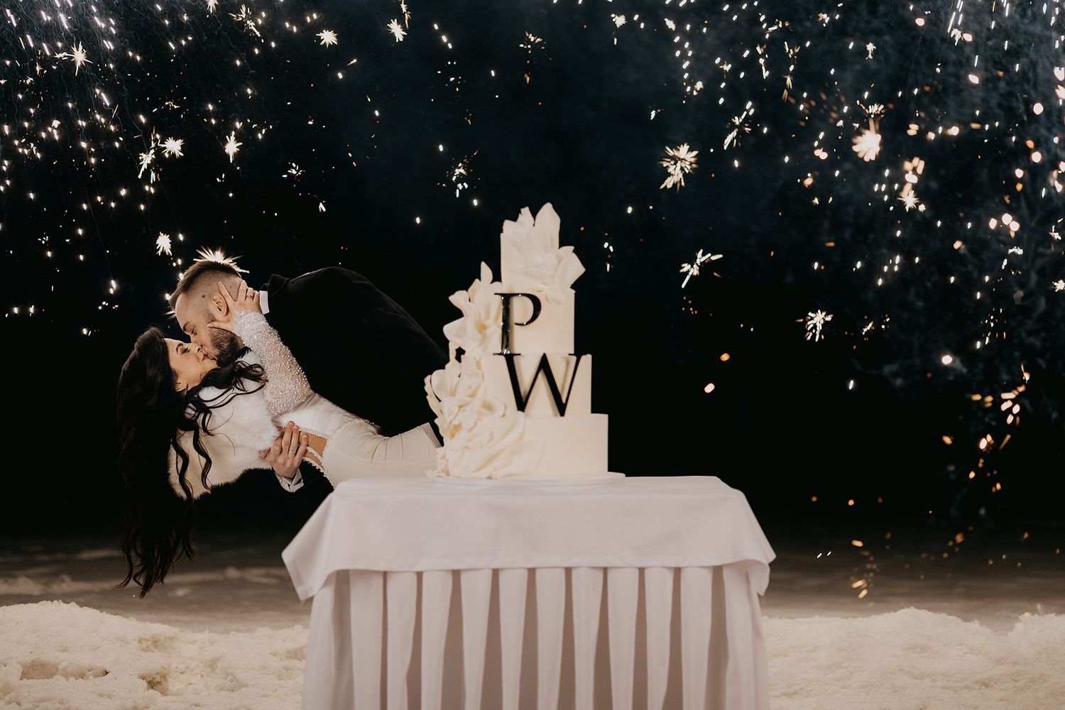 Bride and groom share a romantic dip kiss beside a white wedding cake with PW monogram amid golden sparks.