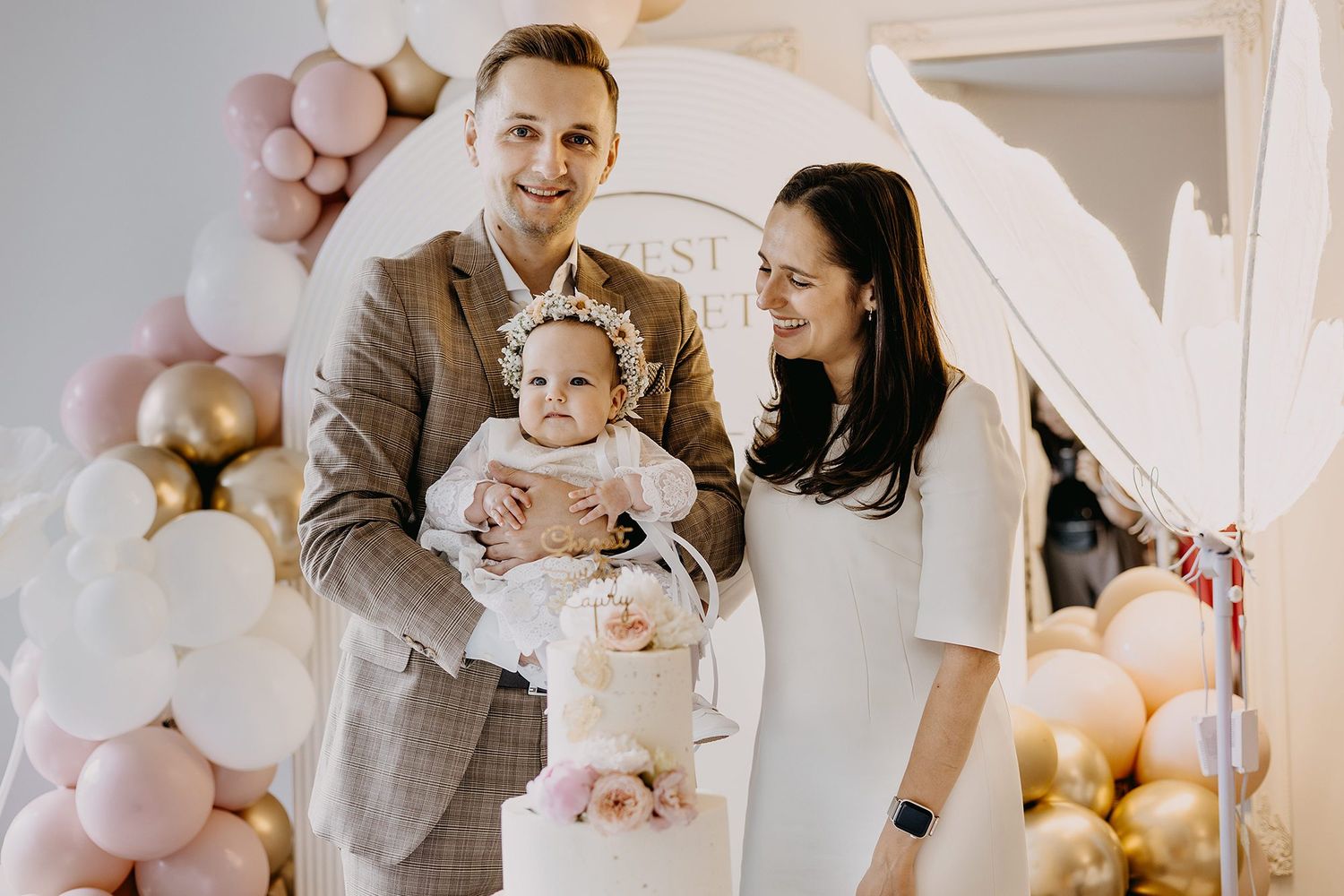 Smiling parents presenting their baby girl at a pink and gold balloon-decorated christening celebration.