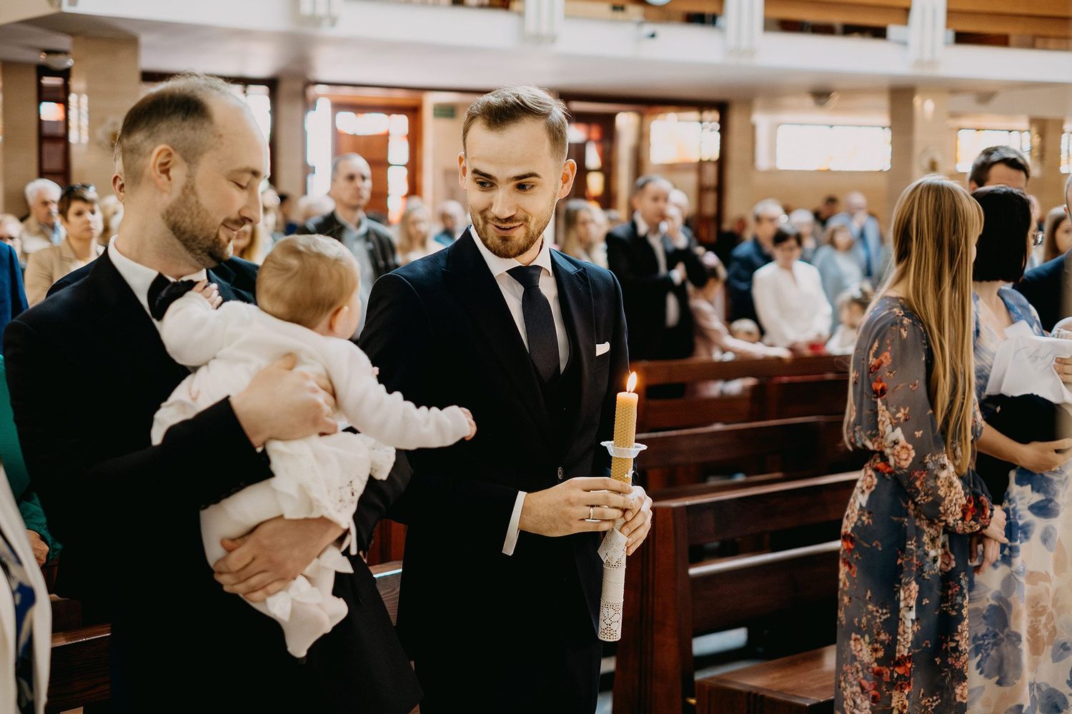 Two men in dark suits hold baby and baptismal candle during church christening ceremony.