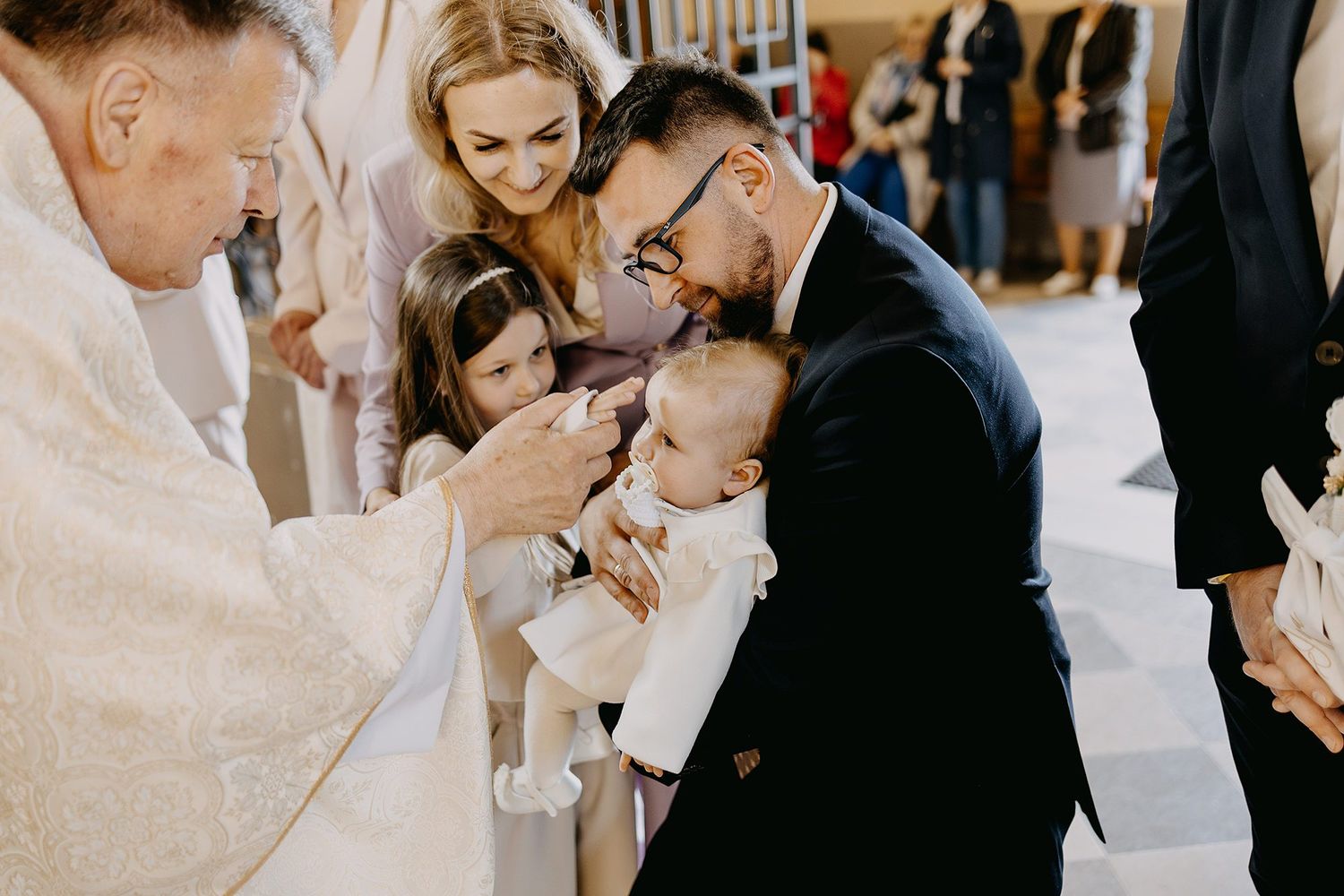 A priest performs a baptism as a father holds his baby with family gathered in church.