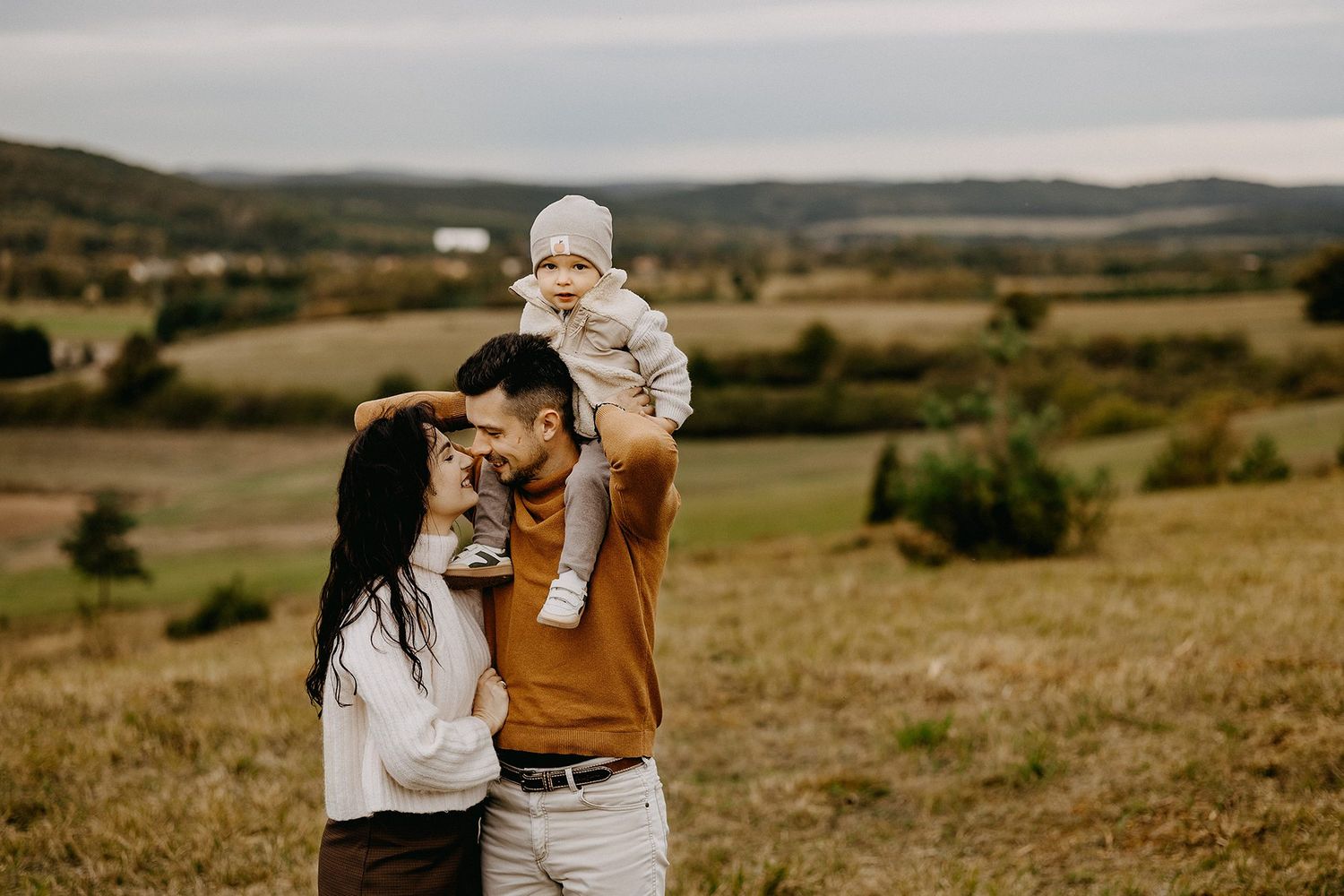 Young couple smiling with baby on shoulders in autumn countryside landscape.
