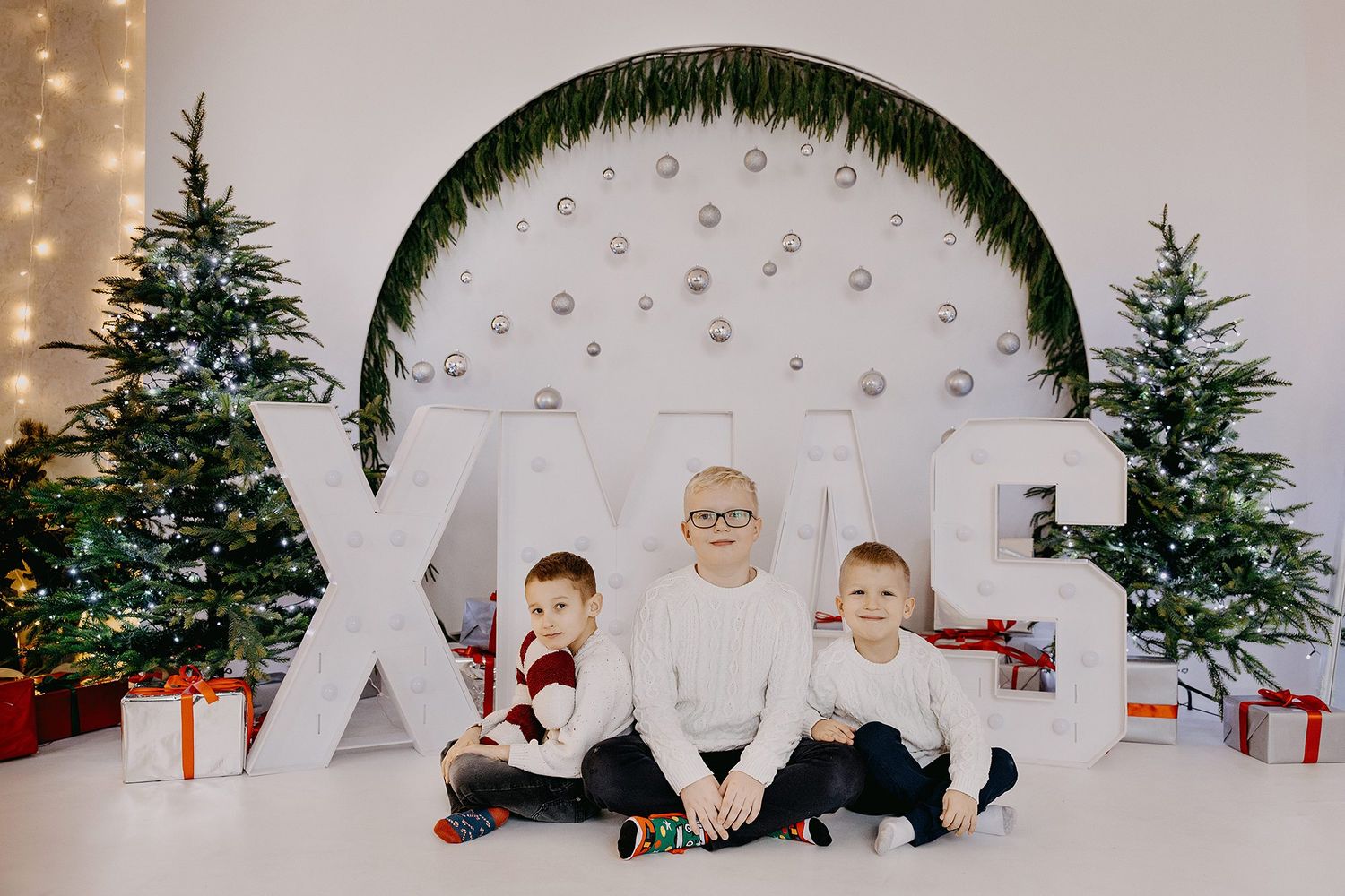 Three boys sit in front of large XMAS letters surrounded by Christmas trees and colorful wrapped presents.