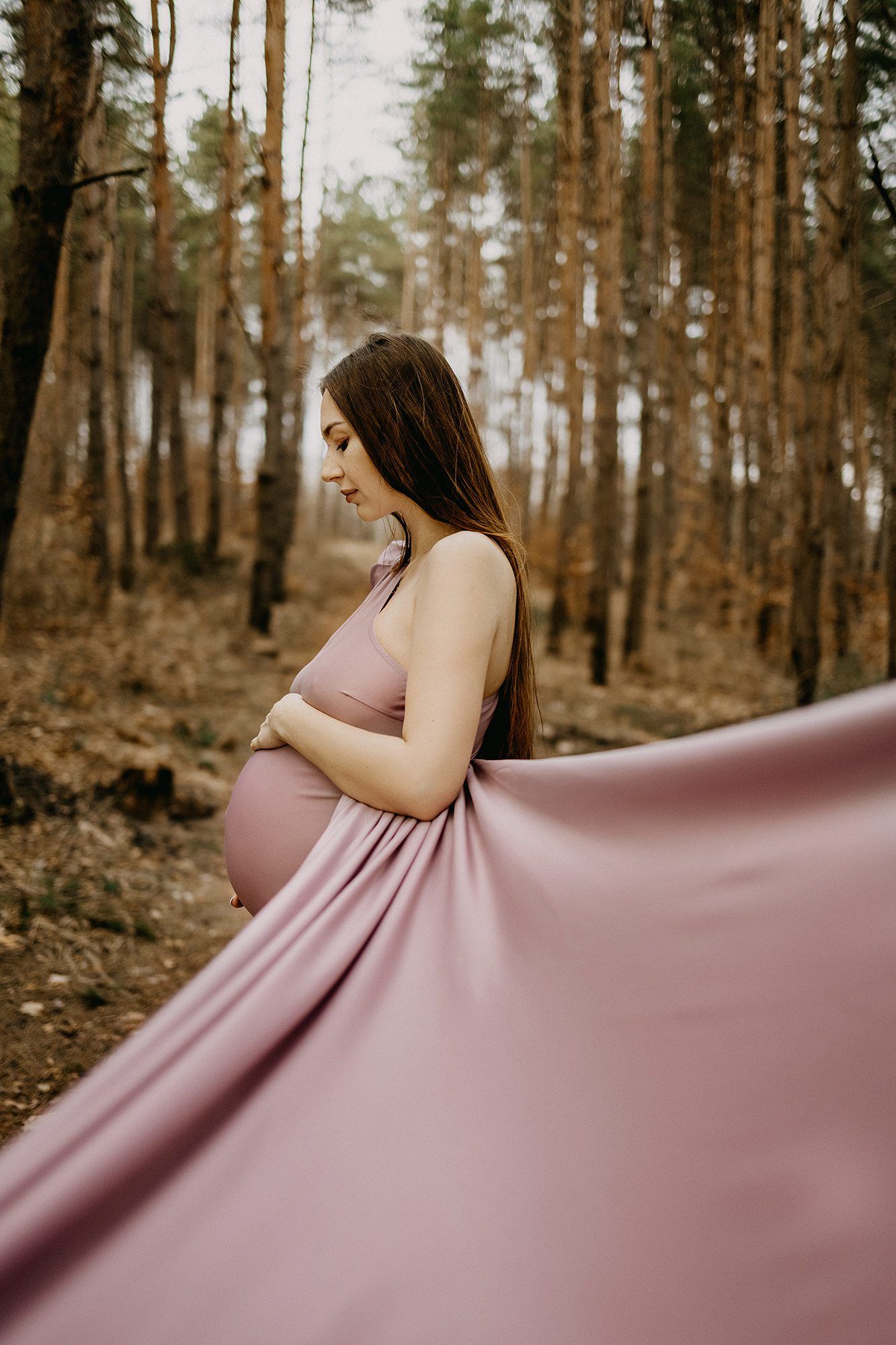 Elegant pregnant woman in flowing pink dress holding baby bump in a serene autumn pine forest.