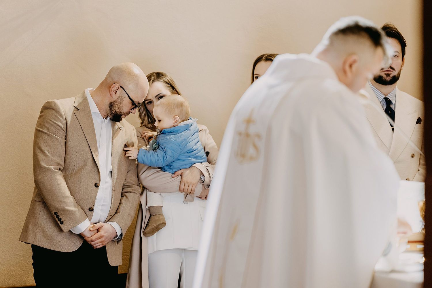 Parents bowing heads in prayer during baptism ceremony, mother holding baby in blue outfit near priest.
