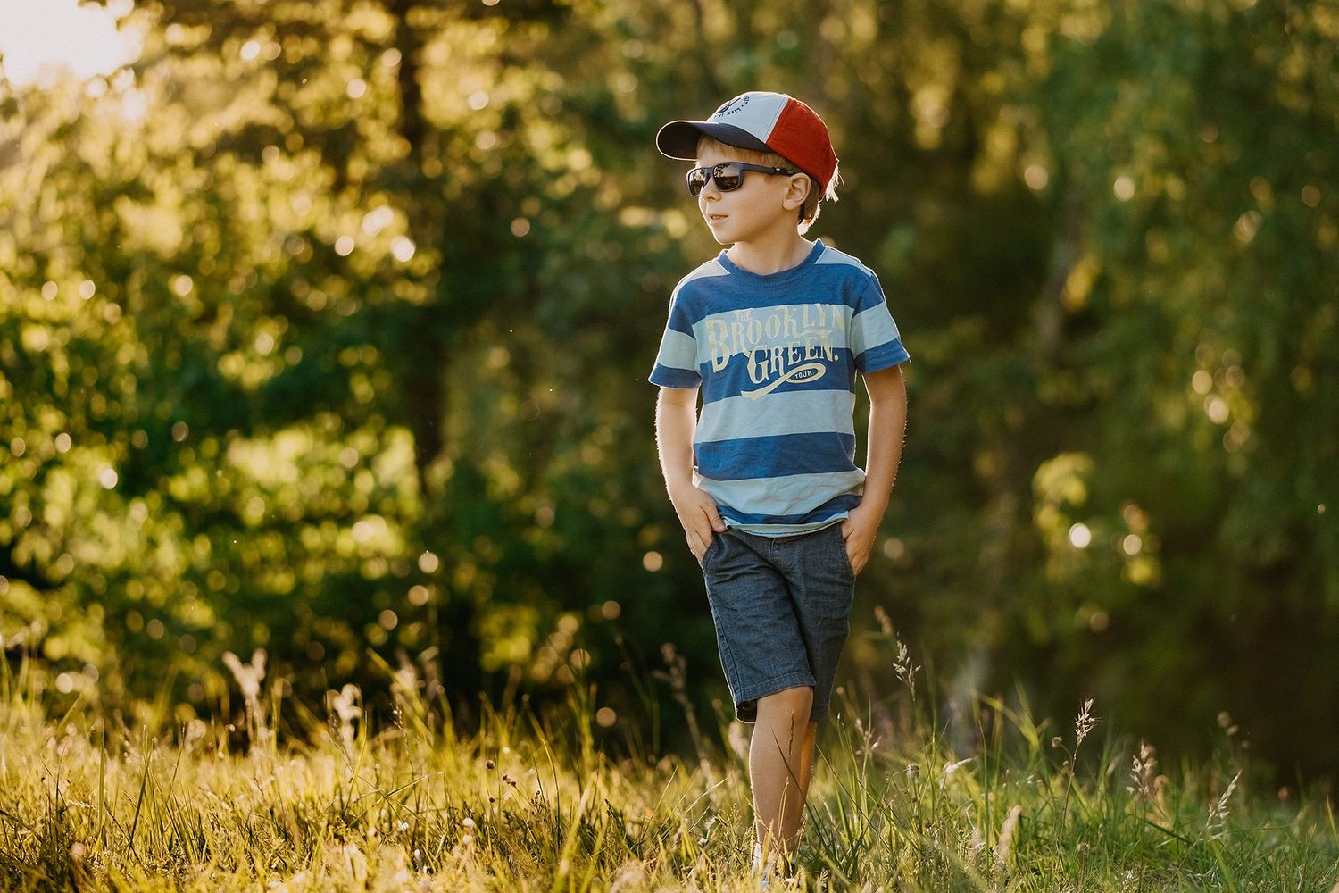 Cute young boy wearing sunglasses and a red cap standing outdoors in a sunny green park.