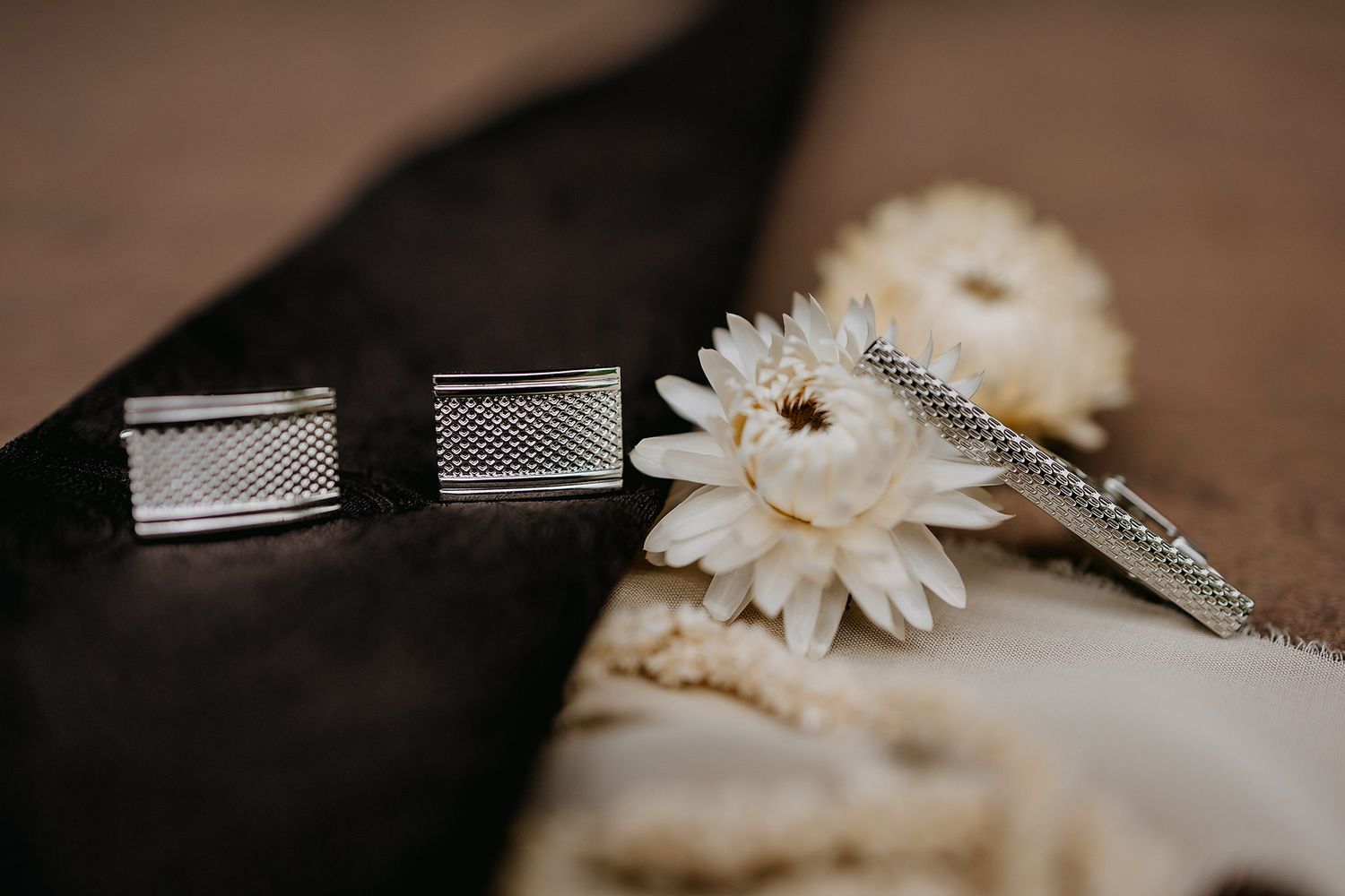 Silver rectangular cufflinks and tie clip displayed beside white chrysanthemum flower on dark fabric.