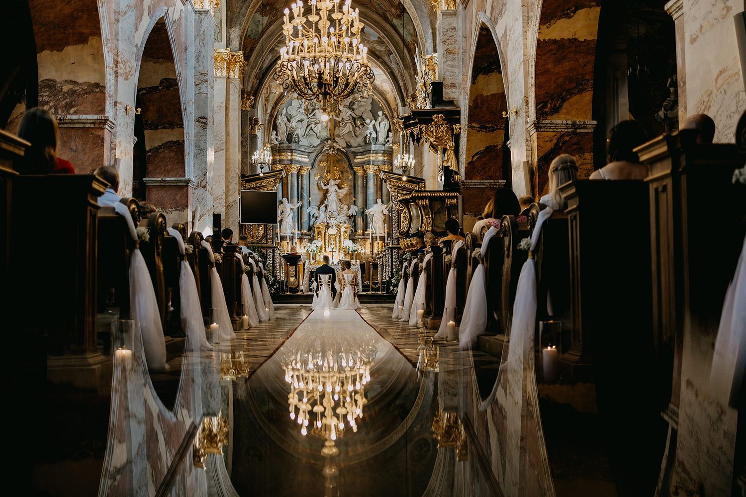 Elegant church wedding ceremony with chandeliers illuminating a long aisle as bride walks toward the altar.