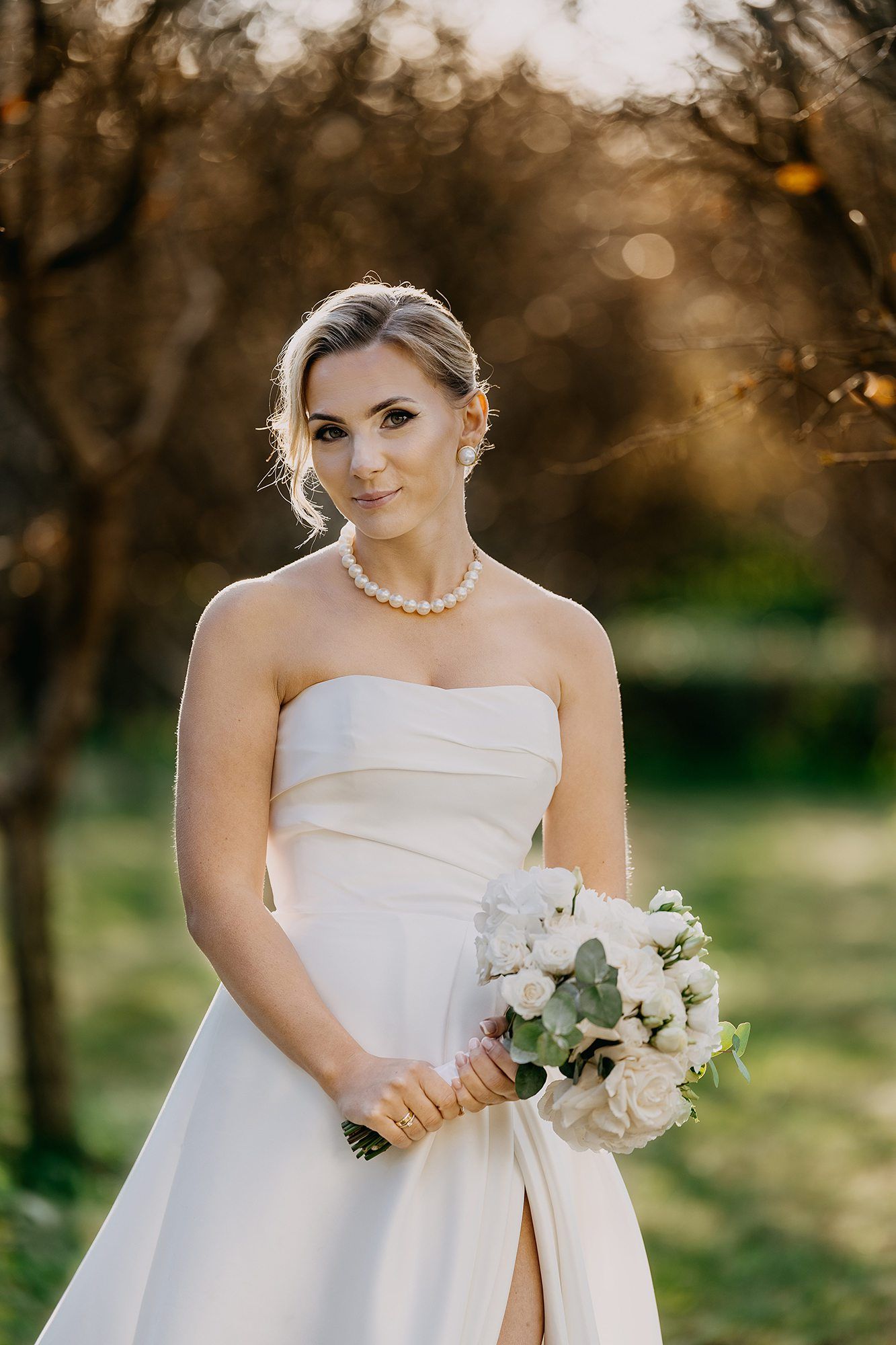 Elegant bride in strapless white gown holding white rose bouquet with pearl necklace in autumn garden.