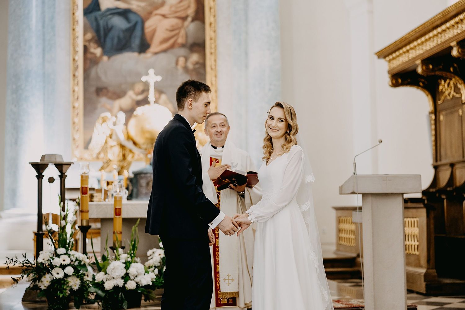 Couple exchanging vows before a priest at a beautifully decorated church altar with floral arrangements.