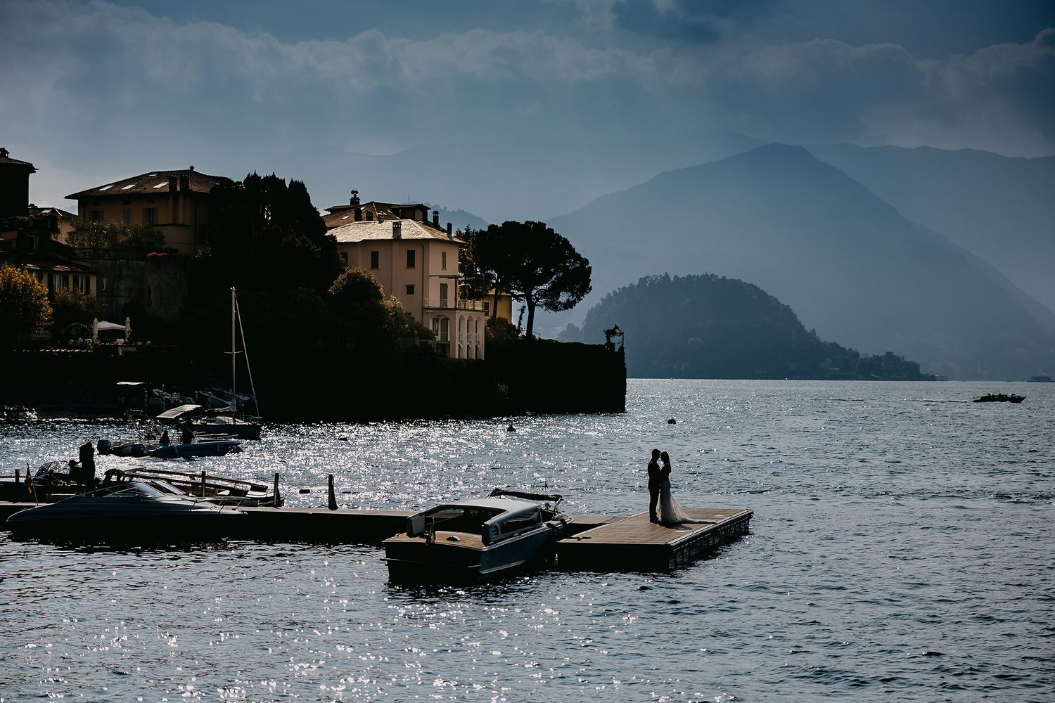 Lone wedding couple stands on a small dock surrounded by shimmering lake water and mountains.