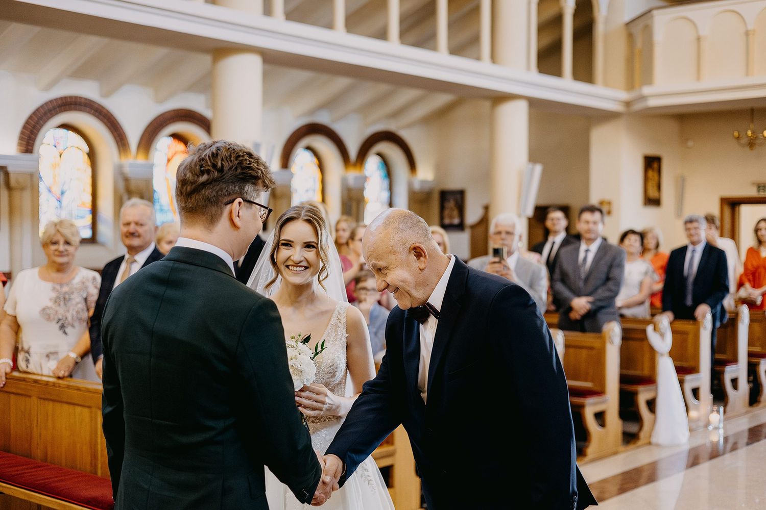 Groom shakes hands with elderly guest while smiling bride stands beside him in church.