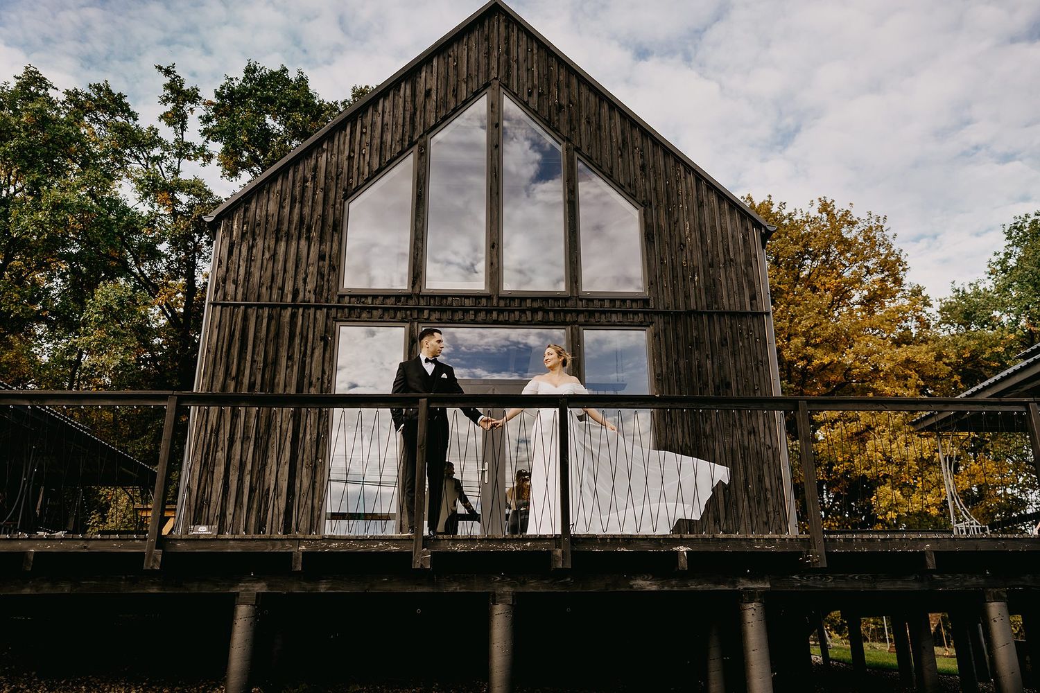 Bride and groom standing on a rustic wooden barn deck surrounded by autumn trees.