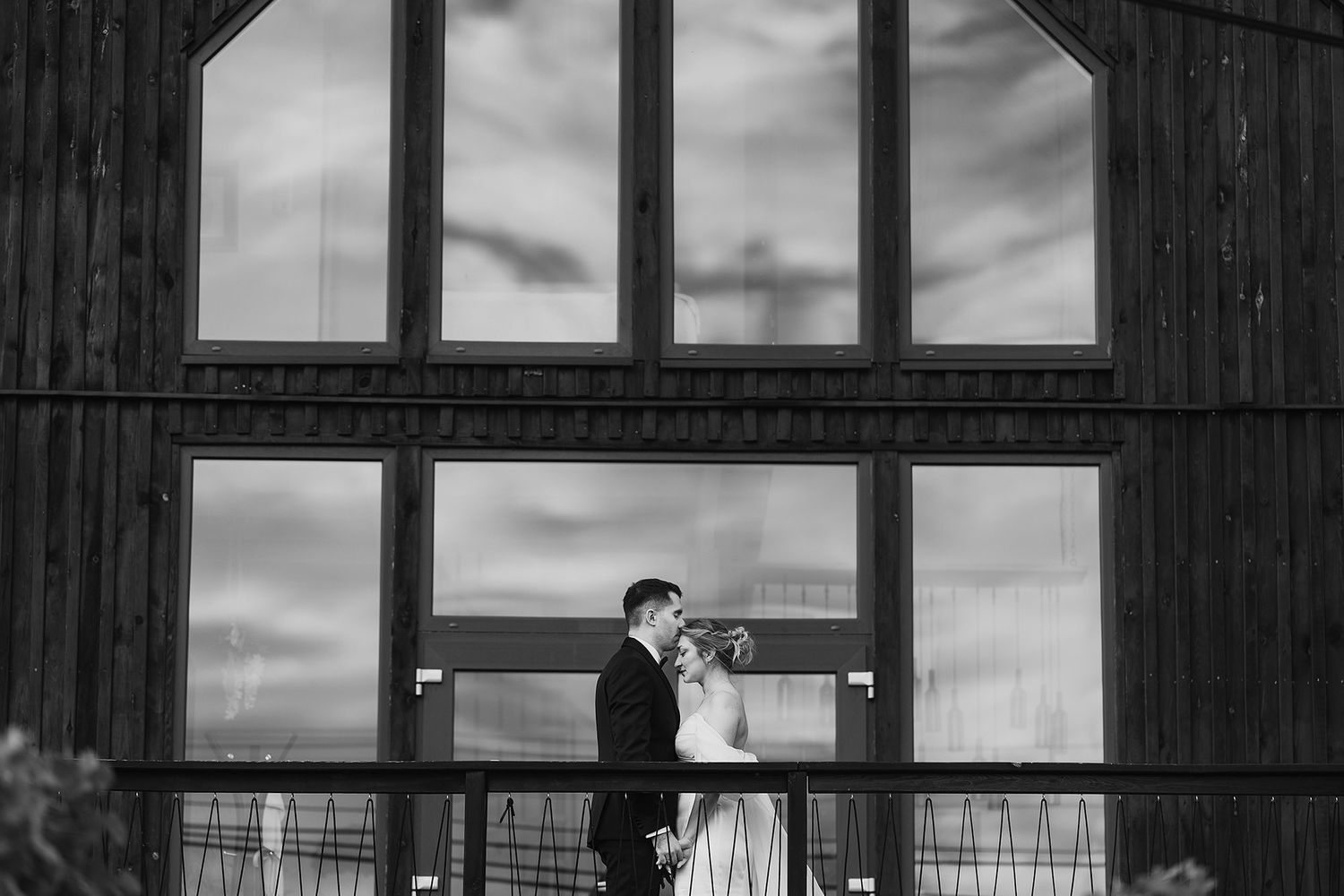 Black and white photo of bride and groom embracing before large barn windows on their wedding day.