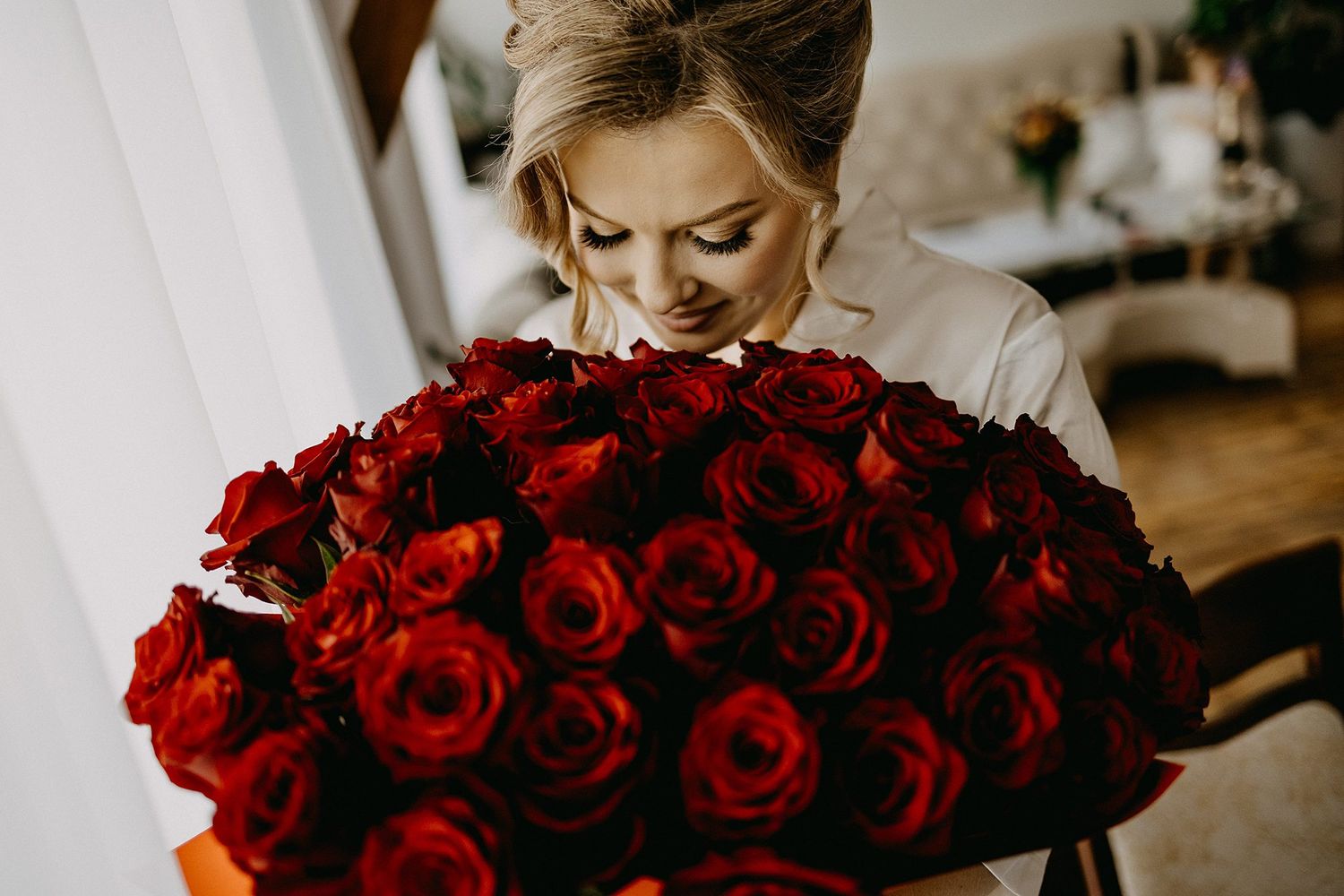 Blonde bride smelling large bouquet of red roses during wedding preparations.