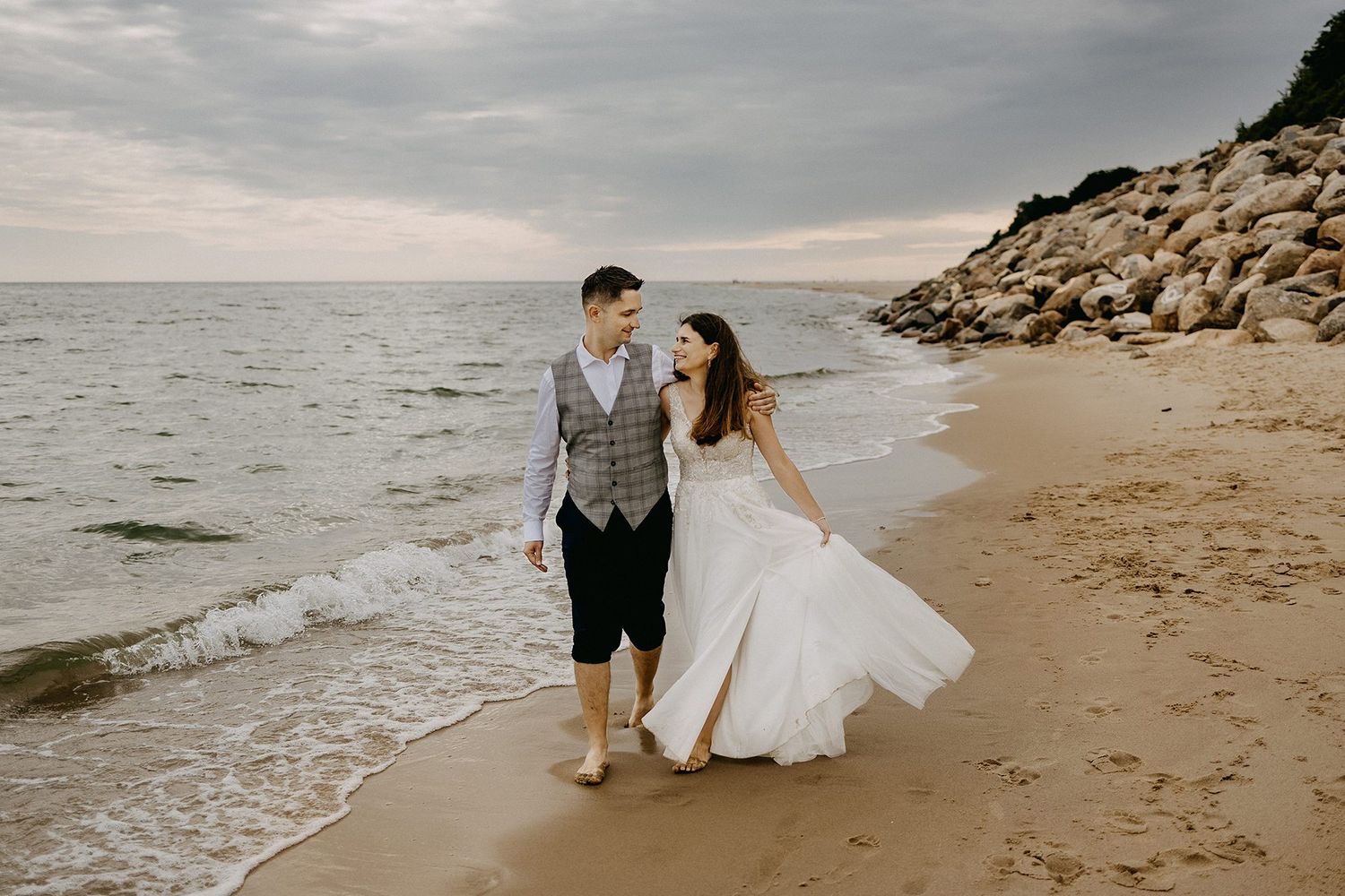 Bride and groom walk barefoot along a sandy beach at sunset with rocky cliffs nearby.
