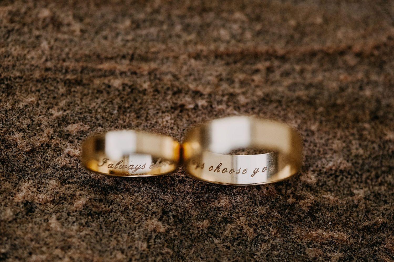 Two gold wedding bands engraved with always and I choose you resting on a textured stone surface.